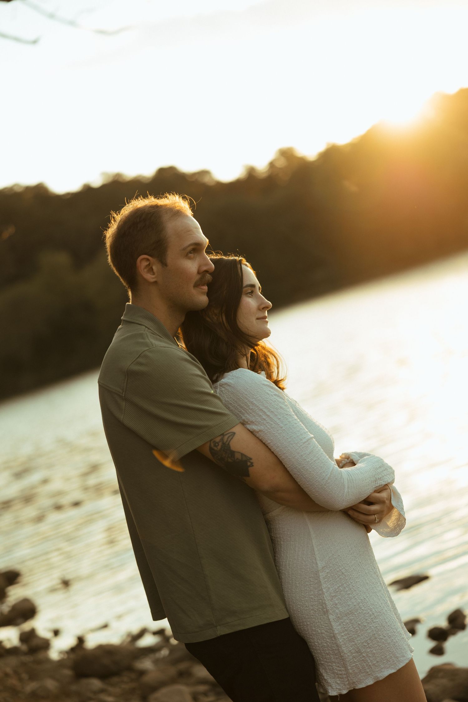 Couple shares tender moment along rocky lakeshore during golden hour sunset.
