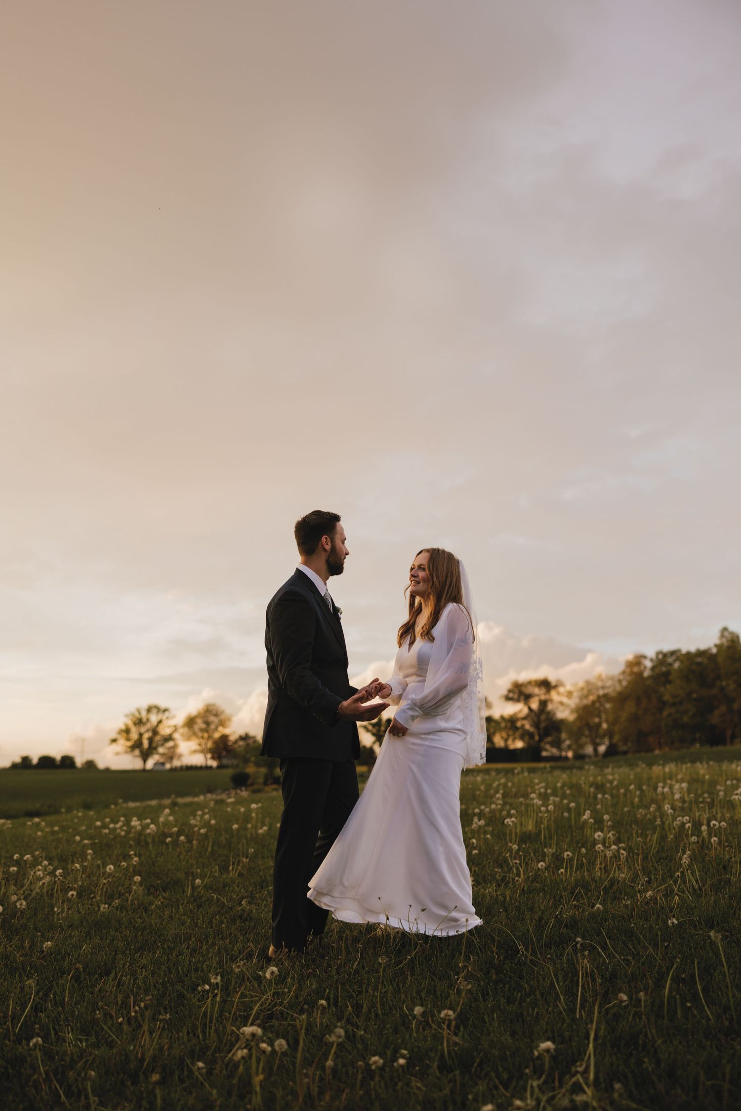 Couple in wedding attire standing in a golden meadow at sunset with a dreamy atmospheric glow.