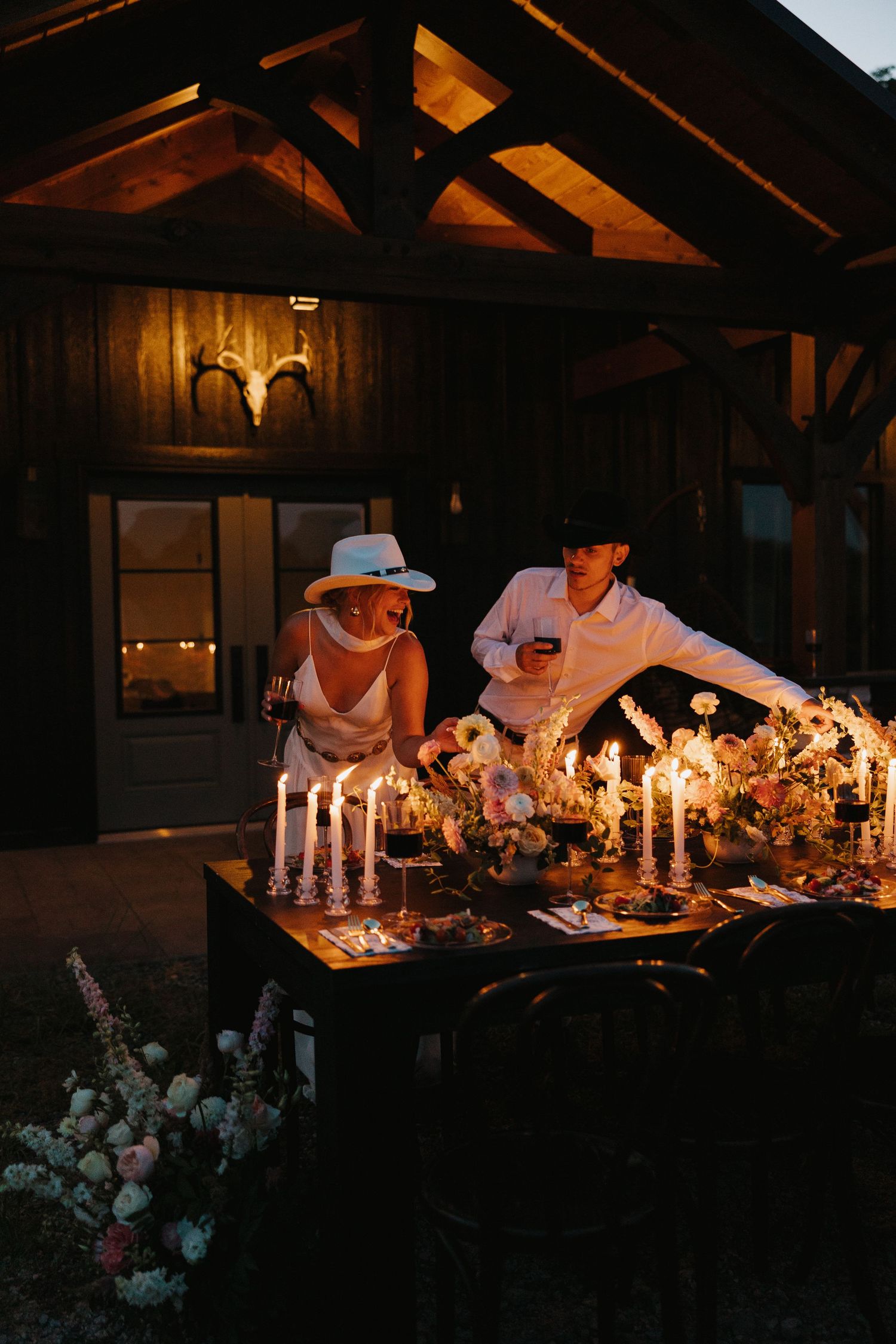 Two people work together to arrange elegant candles and floral decorations on a rustic wooden table at dusk.