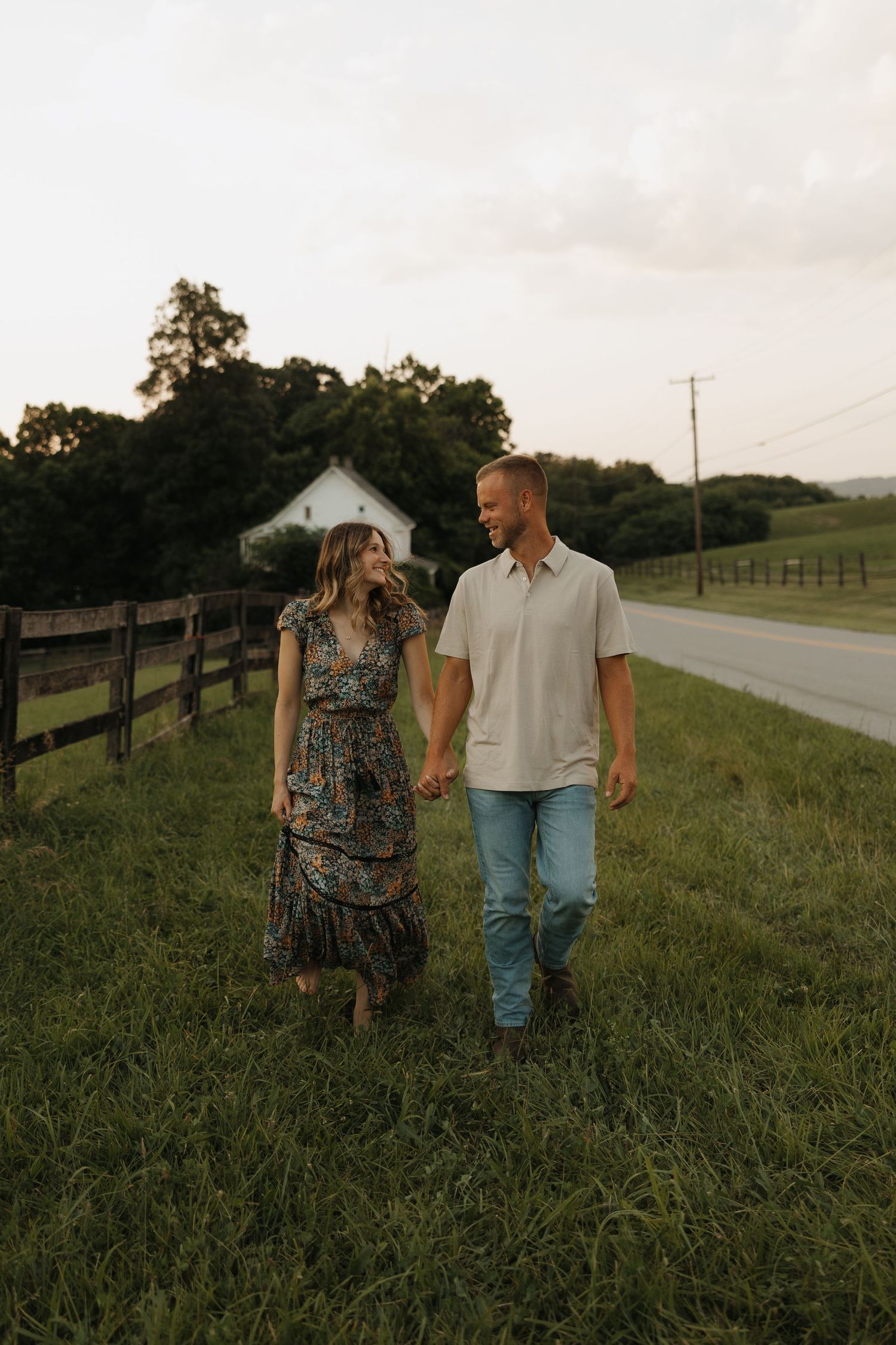 A couple takes a romantic stroll through a grassy field with wooden fences and a country road in the background.