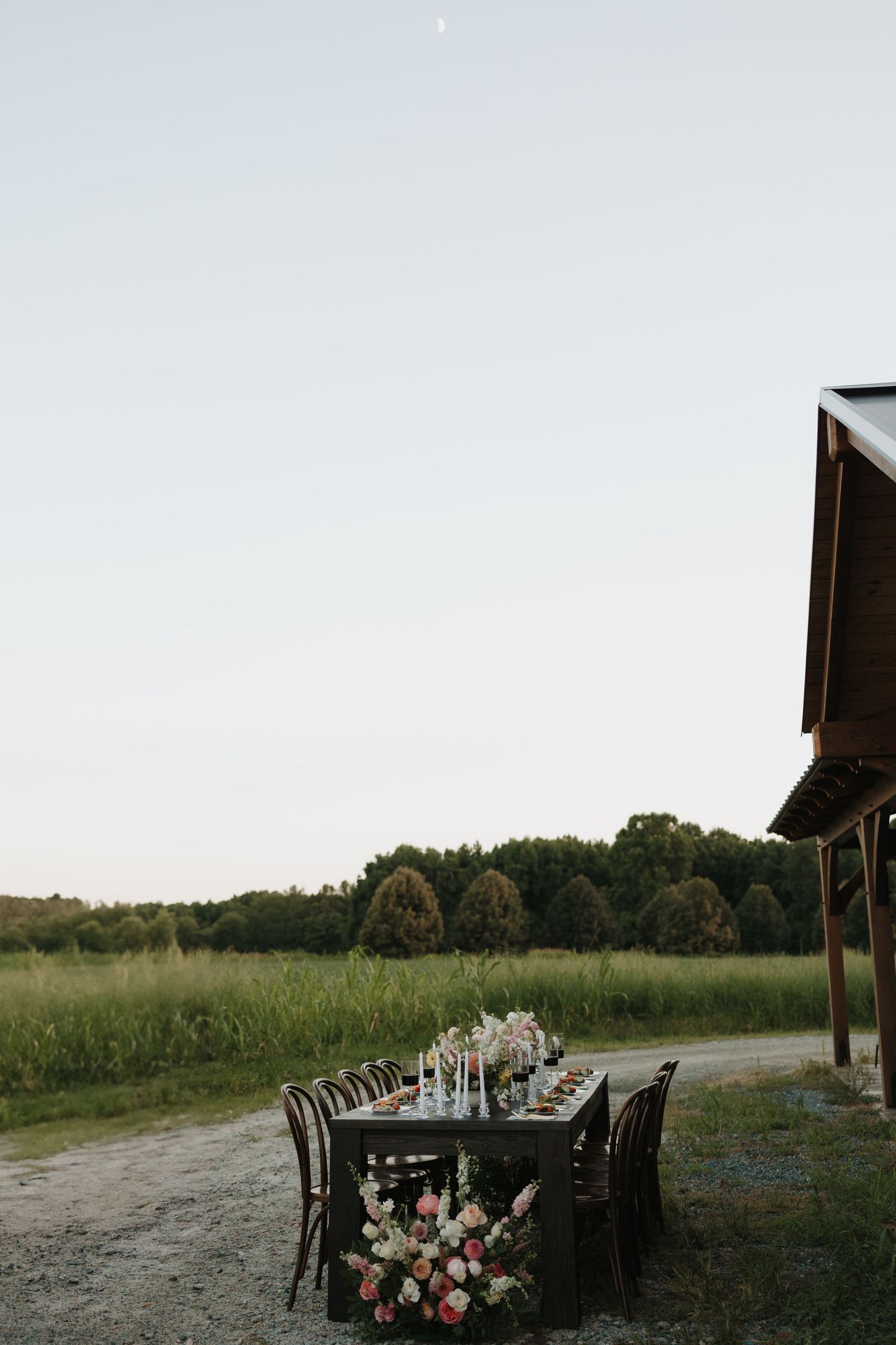 Rustic outdoor dining setup with long wooden tables and chairs arranged in a field at sunset.