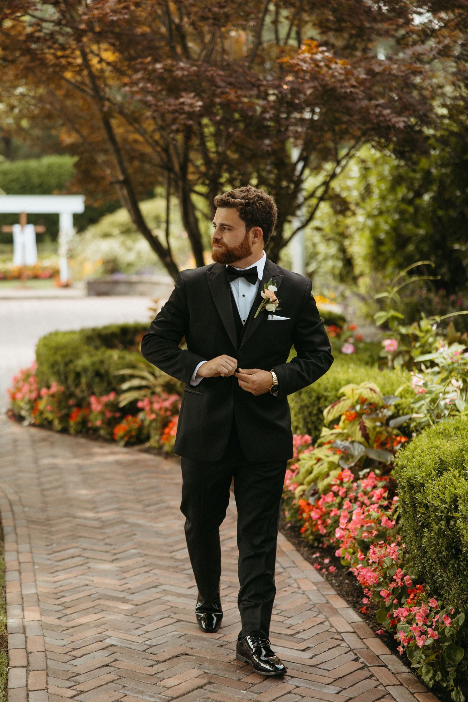 Two people in black suits walk along a brick pathway bordered by colorful flowers and autumn foliage.