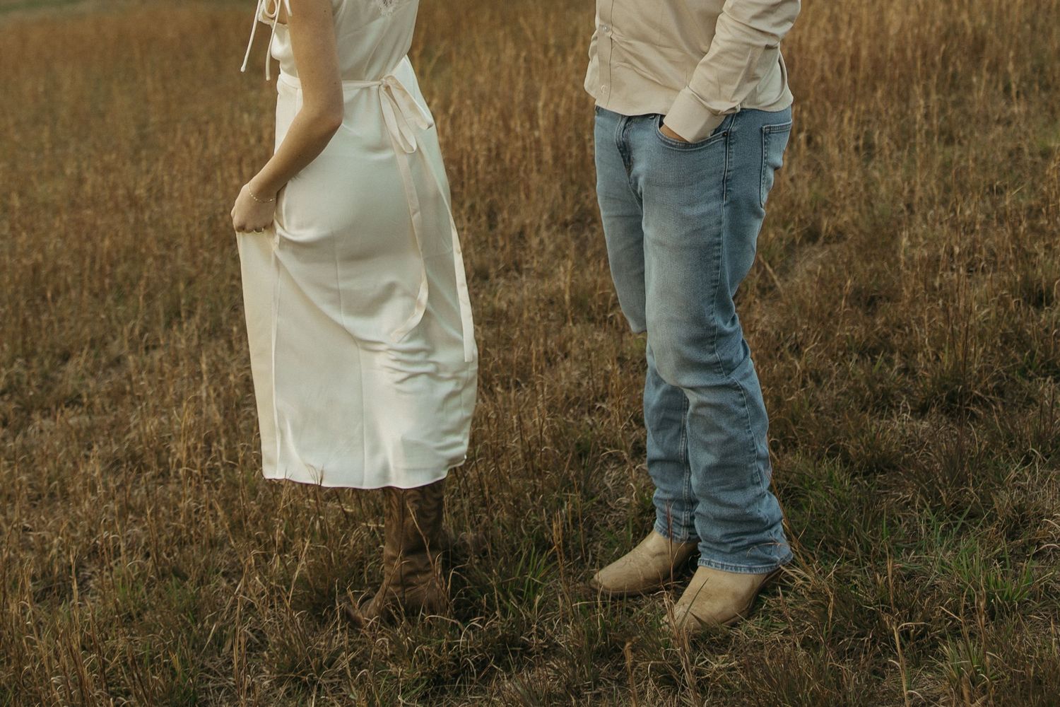 A couple in casual attire stands together in a golden field during sunset.