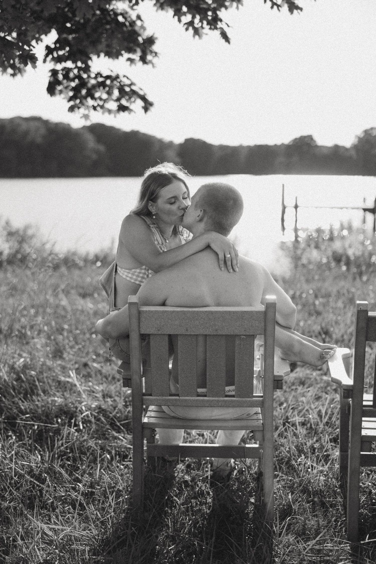 A romantic couple shares intimate moments on a wooden bench by a lake during sunset in black and white photography.