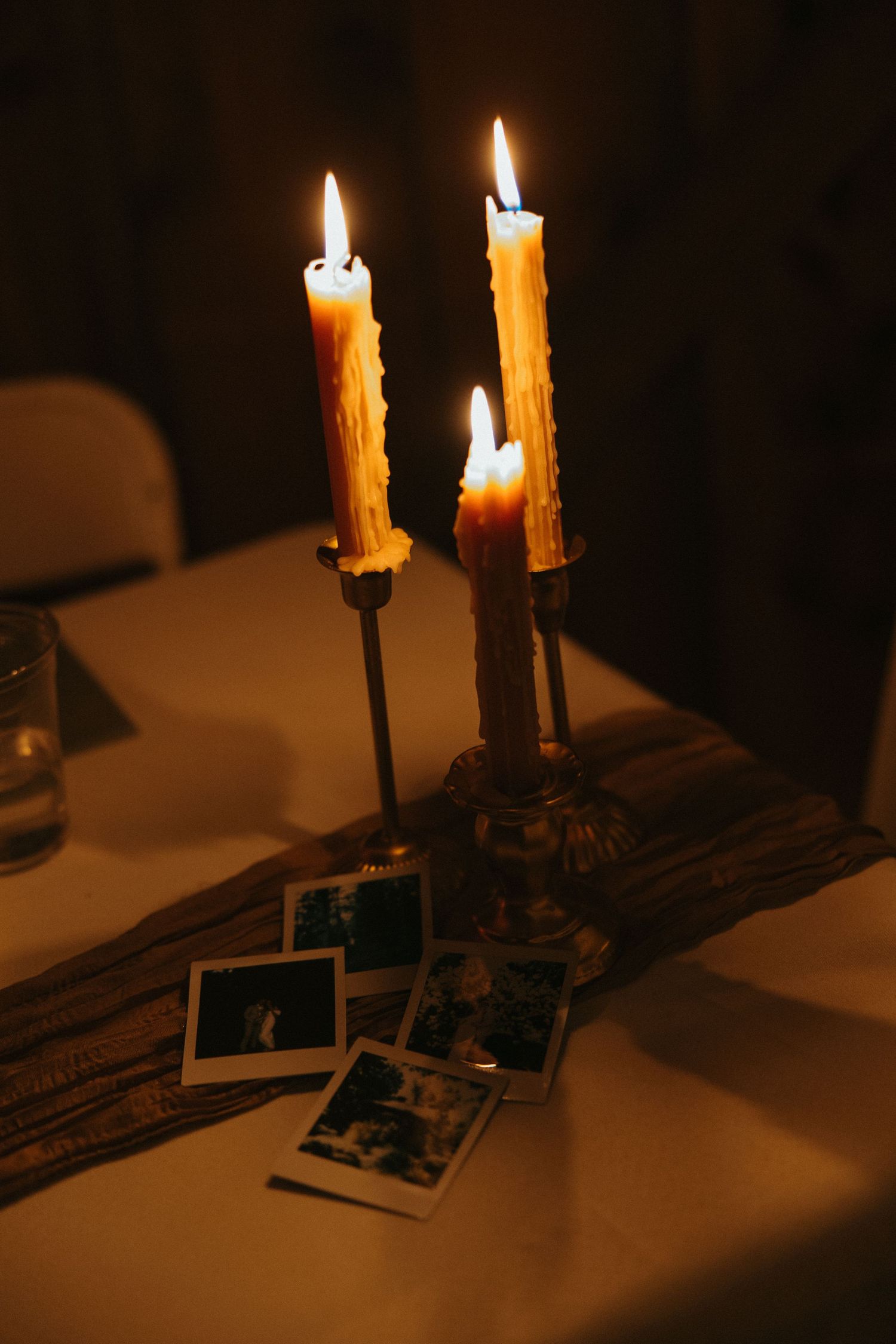 Two lit candles cast a warm glow over scattered photographs on a dark surface in a moody atmospheric scene.