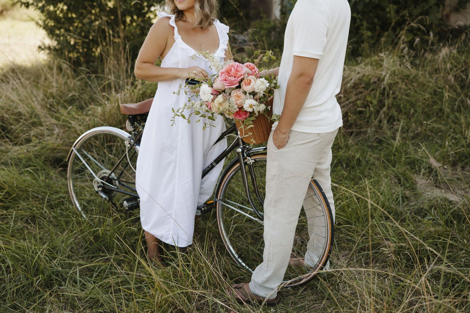 A vintage bicycle adorned with pink and white flowers stands in tall grass next to two people dressed in white.