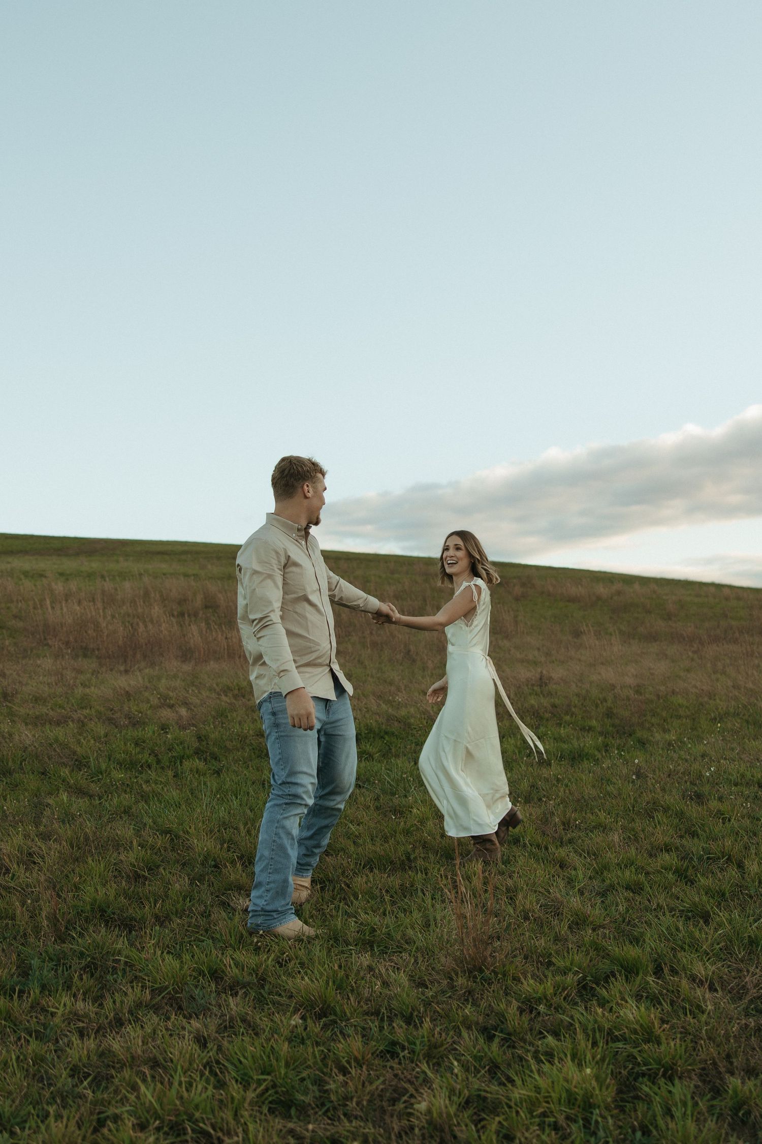 A couple stands together in a grassy field at dusk, with the person in white twirling in a flowing dress.