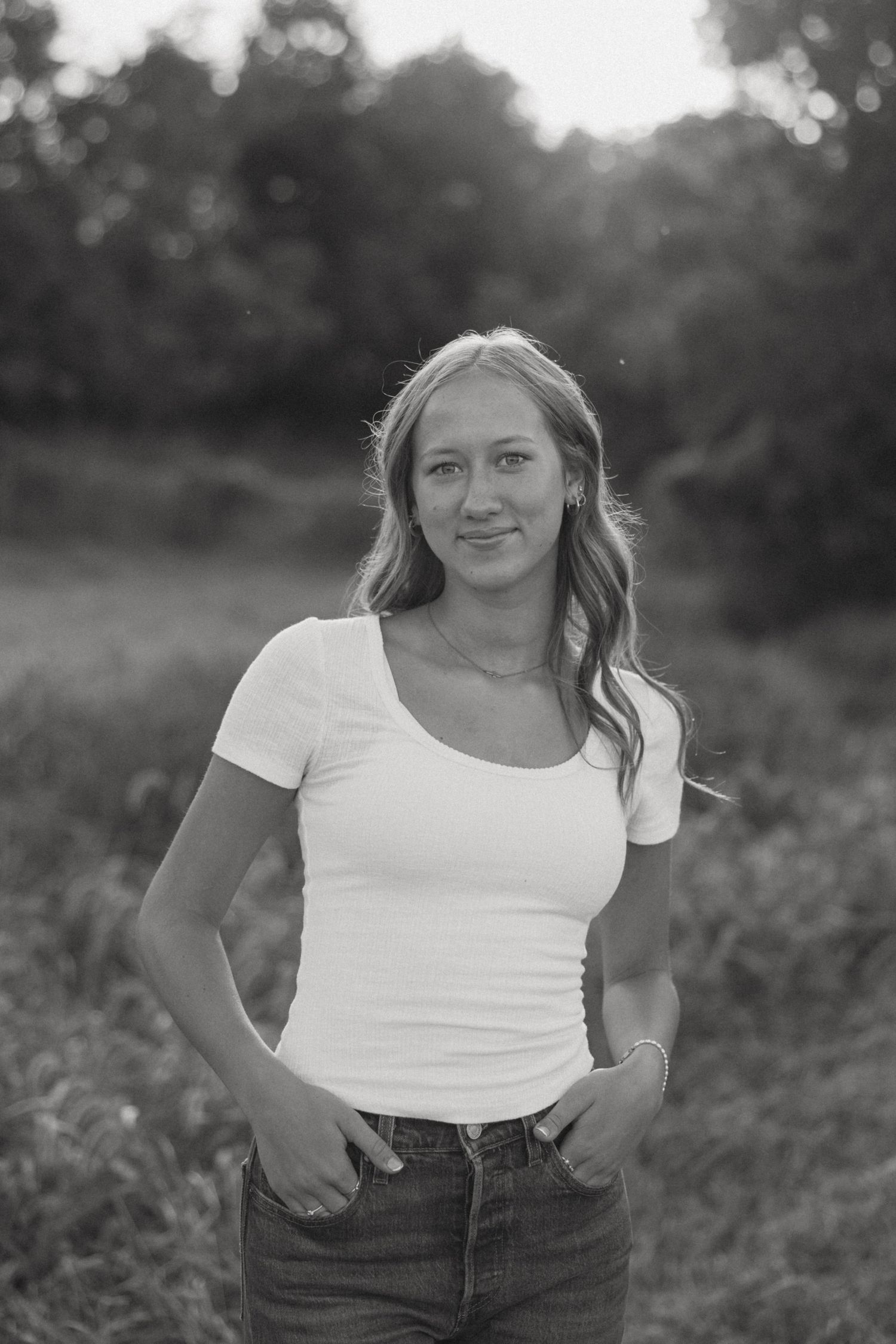 A person in a white t-shirt and jeans poses outdoors in a natural setting with bokeh lighting effects in the background.