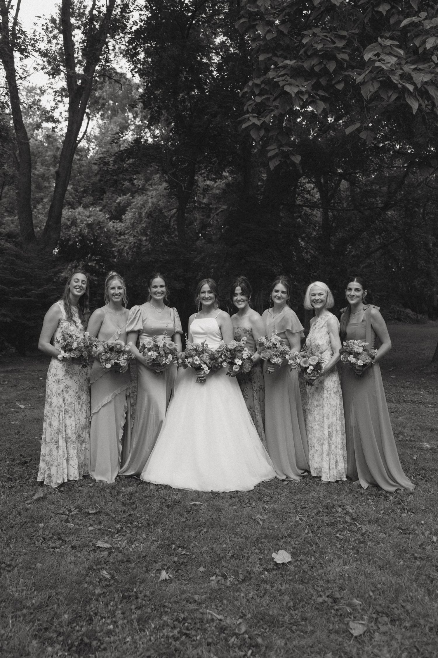 A black and white wedding photo showing a bridal party in mix-matched gowns holding floral bouquets in an outdoor setting.
