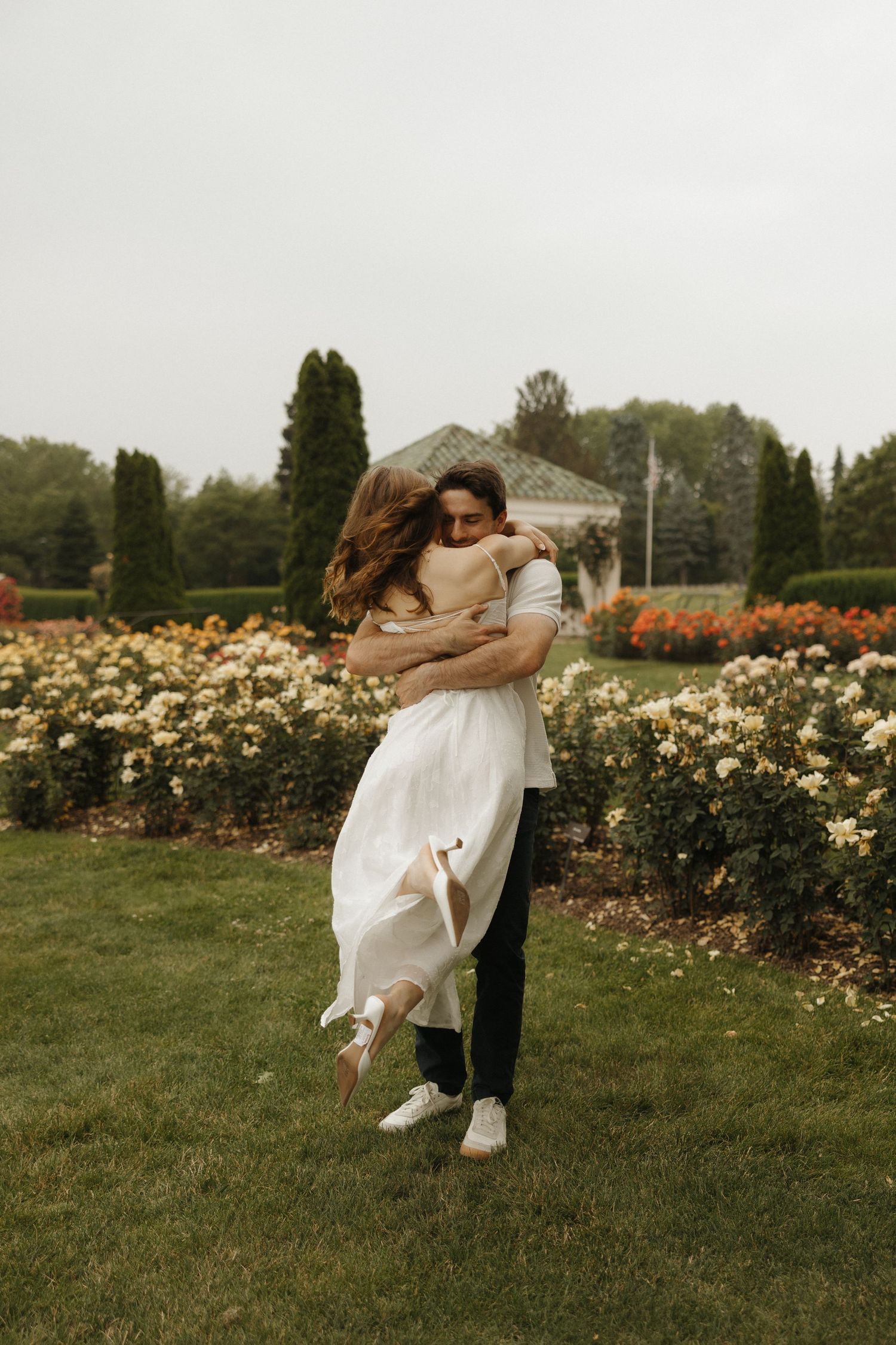 A couple embraces romantically in a lush garden filled with blooming roses on a warm summer day.