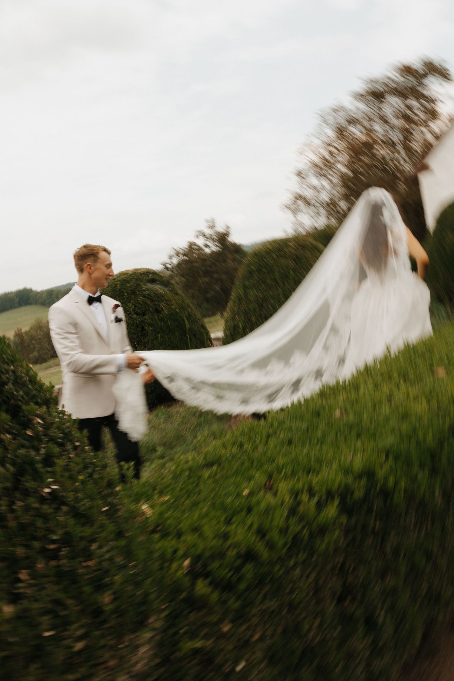 A dramatic motion shot of a white wedding dress train flowing in the wind across a green hillside.
