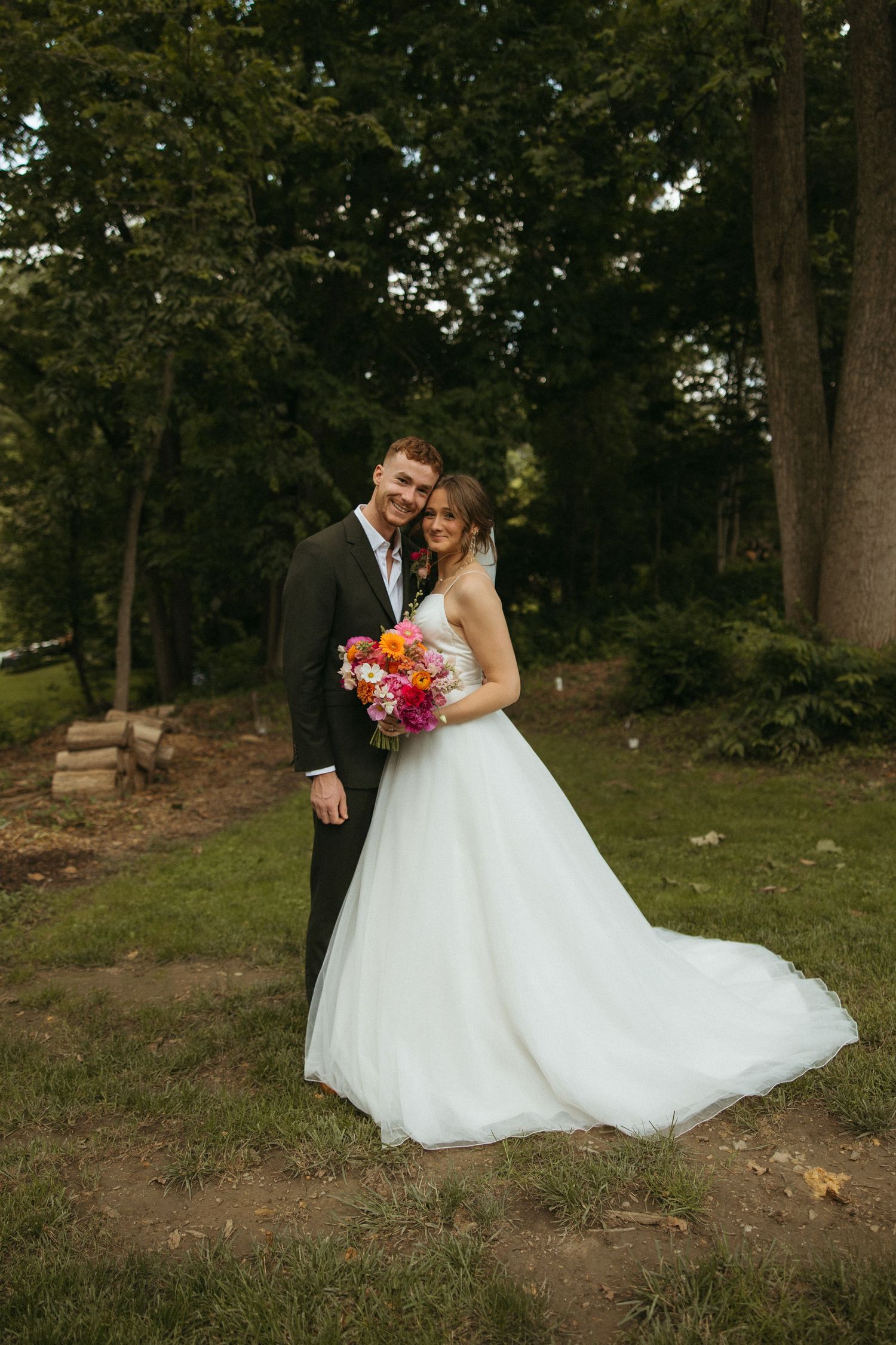 Newlywed couple shares intimate moments in a wooded outdoor setting with bride in flowing white gown and colorful bouquet.