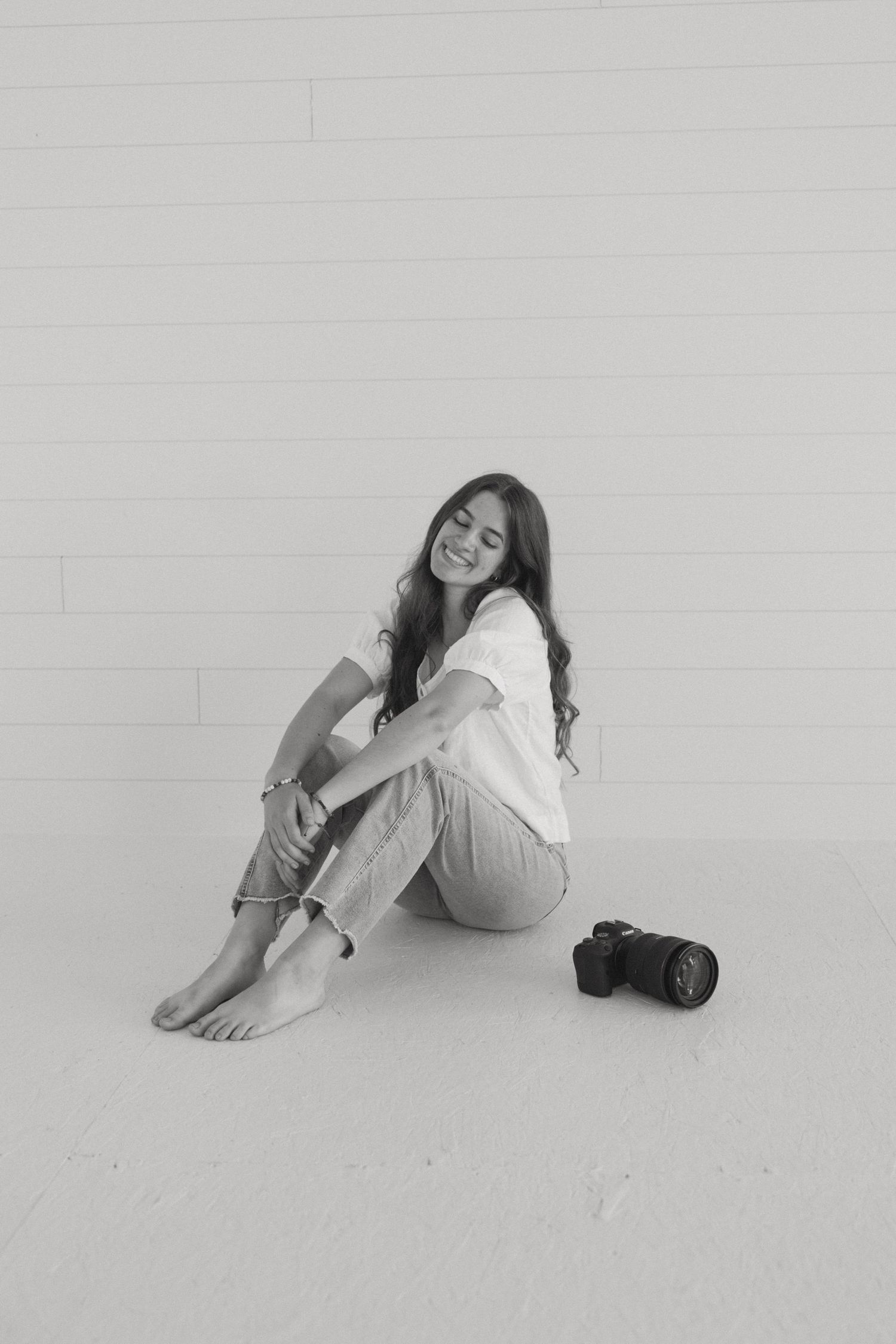 Black and white portrait of someone sitting casually against a wall with a camera nearby.