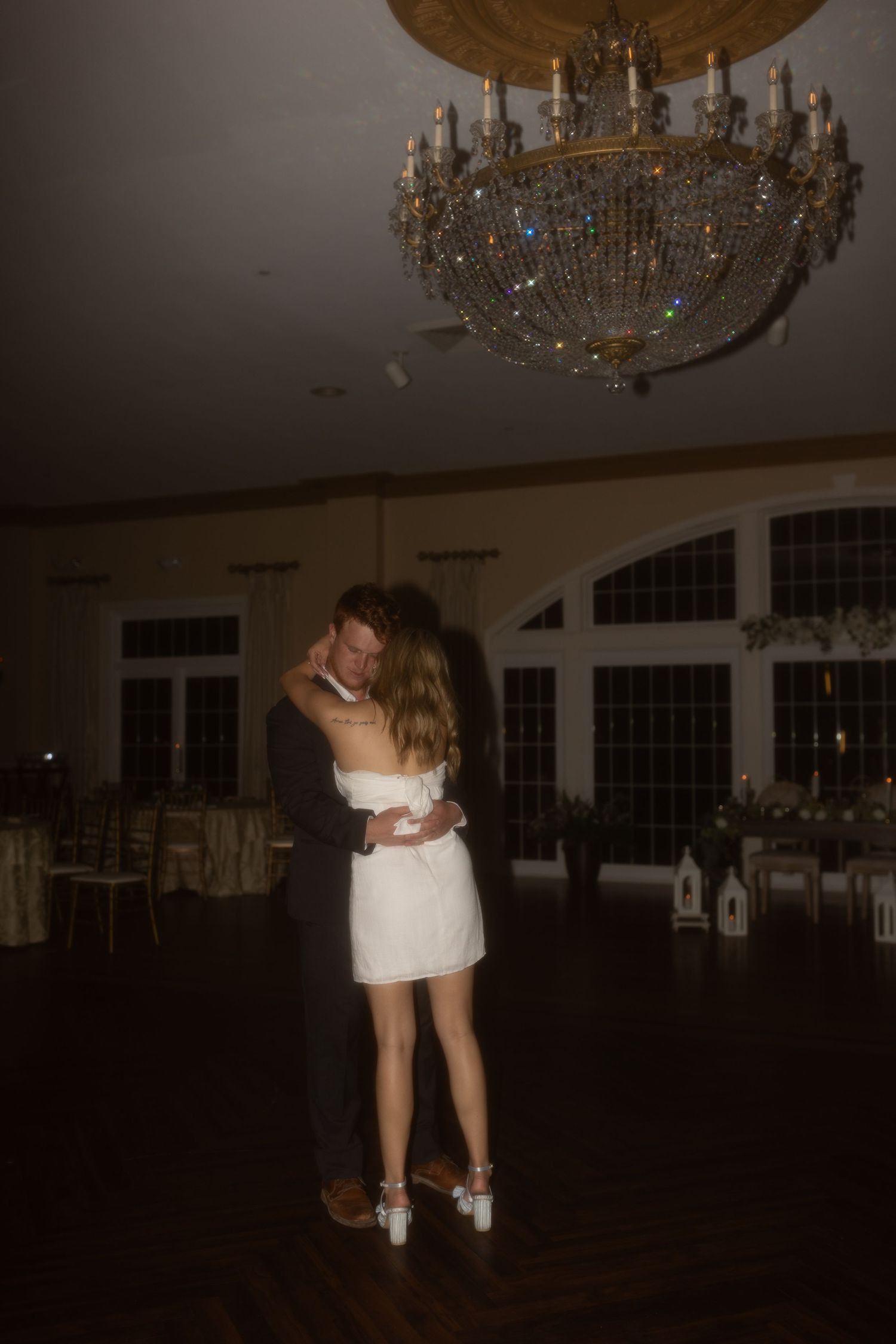A couple shares an intimate dance moment under an ornate crystal chandelier in a dimly lit ballroom.
