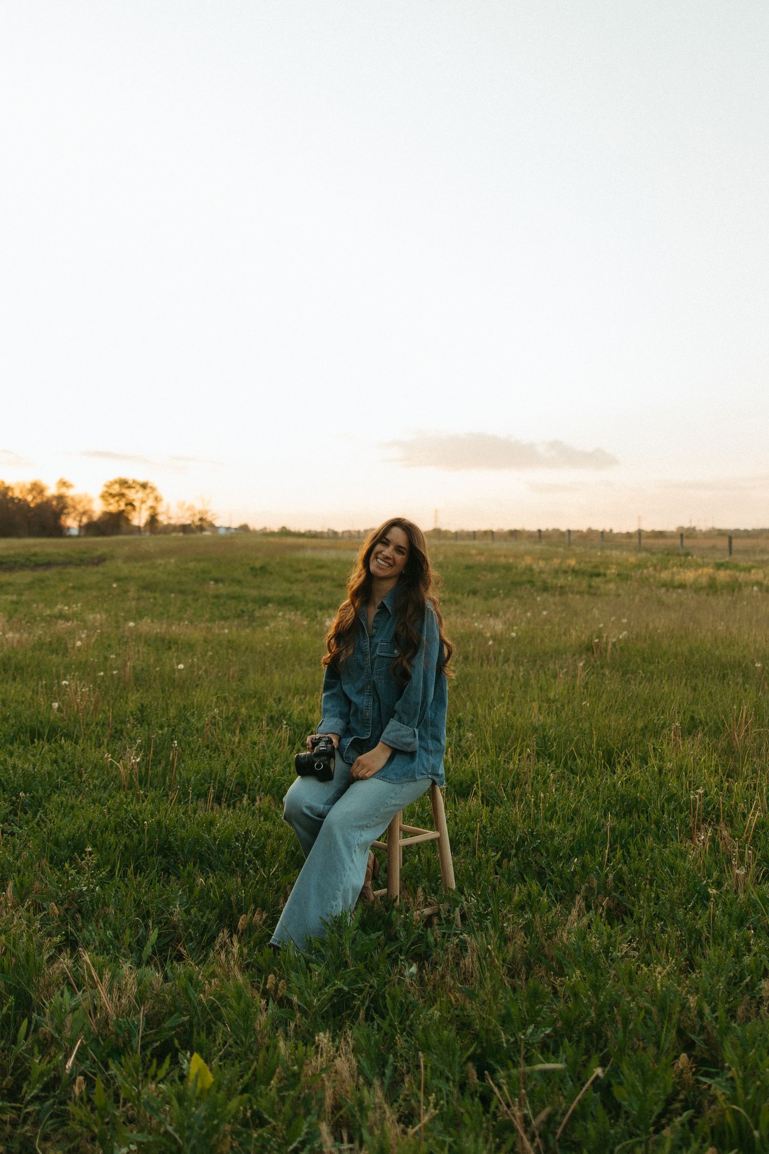 Woman in denim jacket and jeans sitting on wooden stool in green field at sunset holding camera.