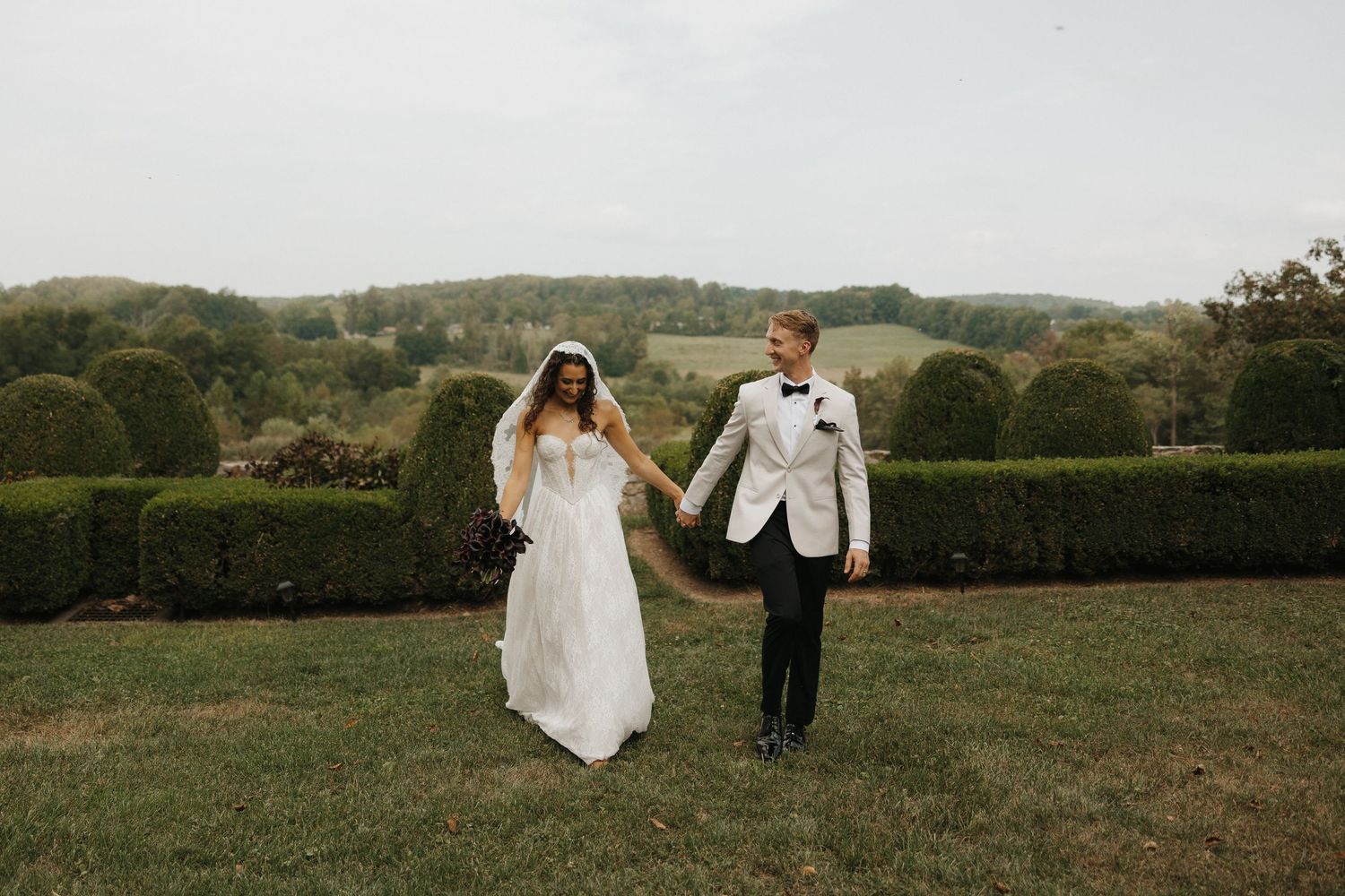 A couple in wedding attire walks through a scenic green field with rolling hills in the background.