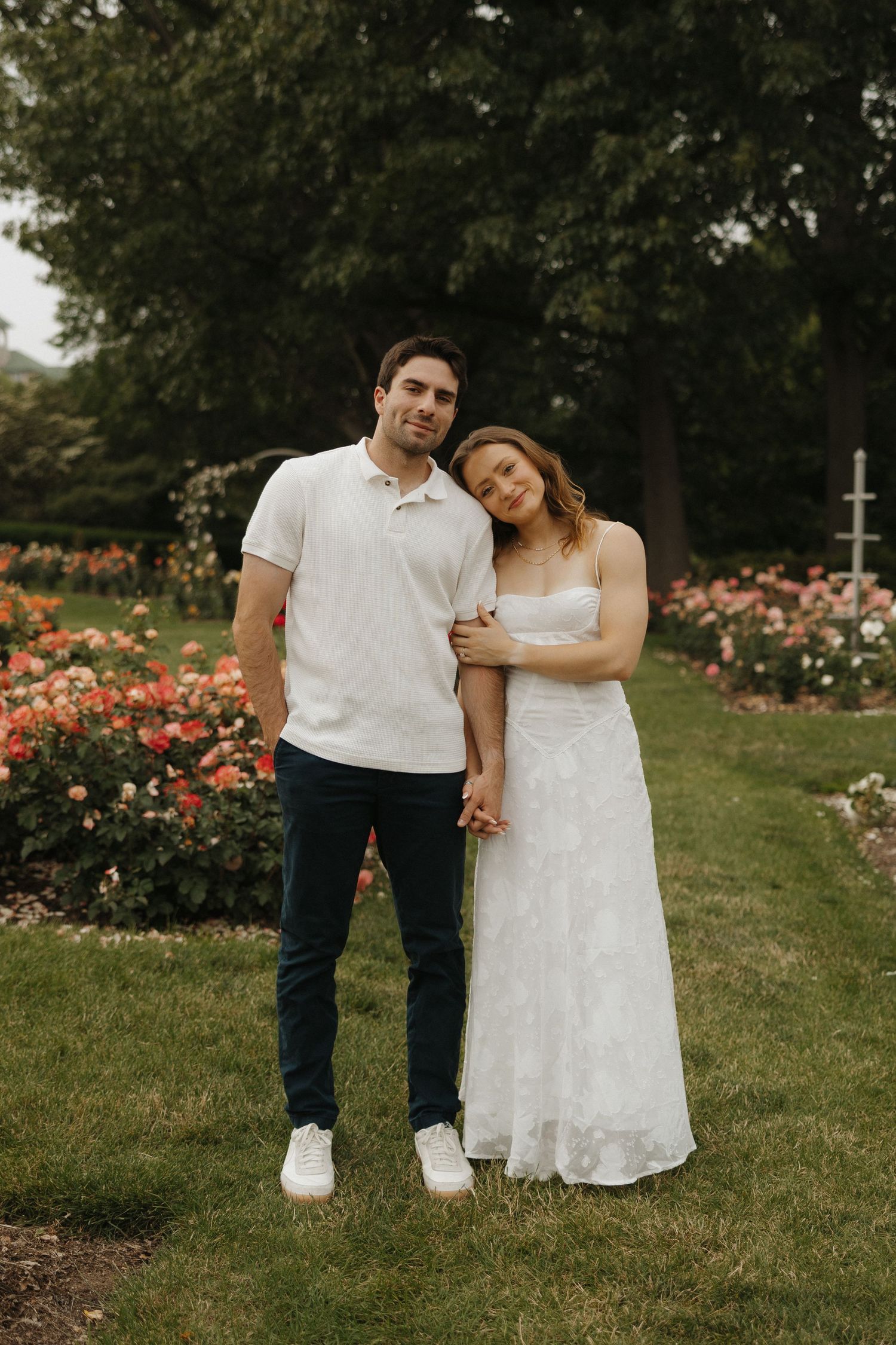 A couple in white and navy outfits poses together in a garden with blooming orange roses in the background.