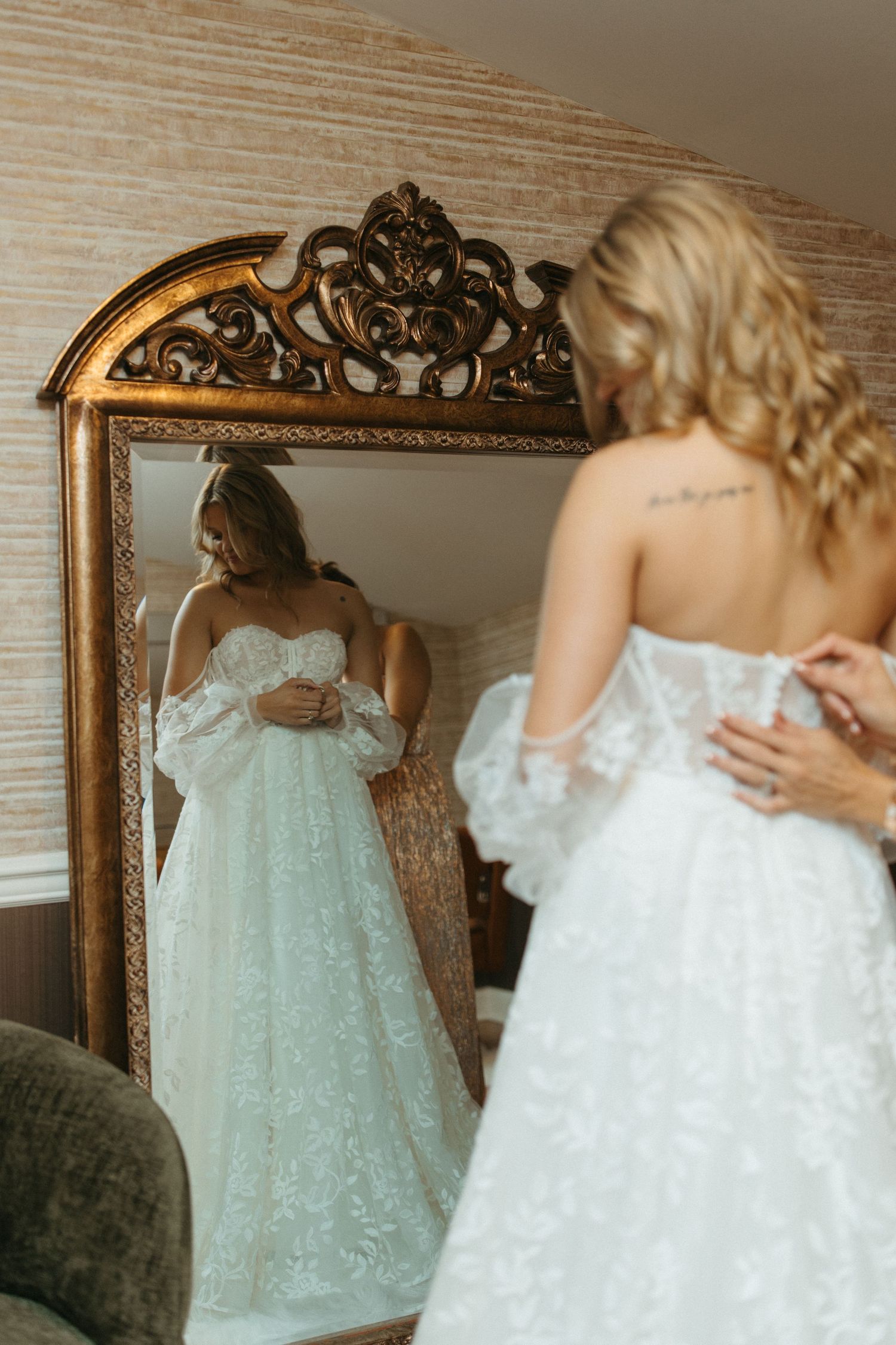 A bride in a white lace wedding gown looks at her reflection in an ornate gold-framed mirror.
