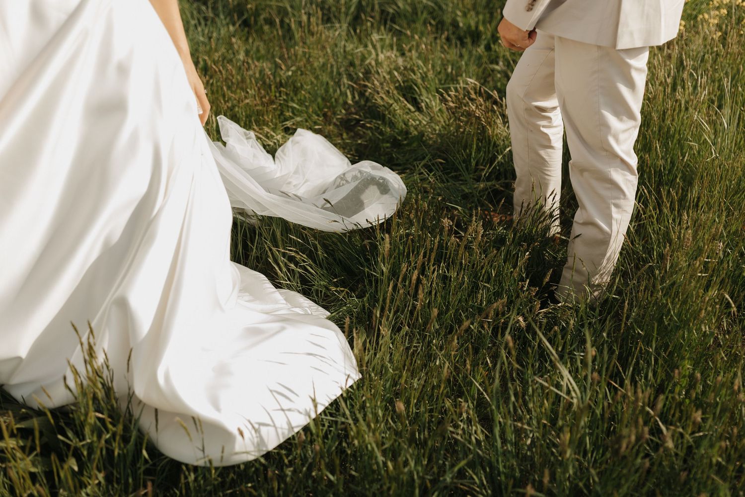 A bride in a flowing white dress walks through tall grass in a rustic outdoor setting.