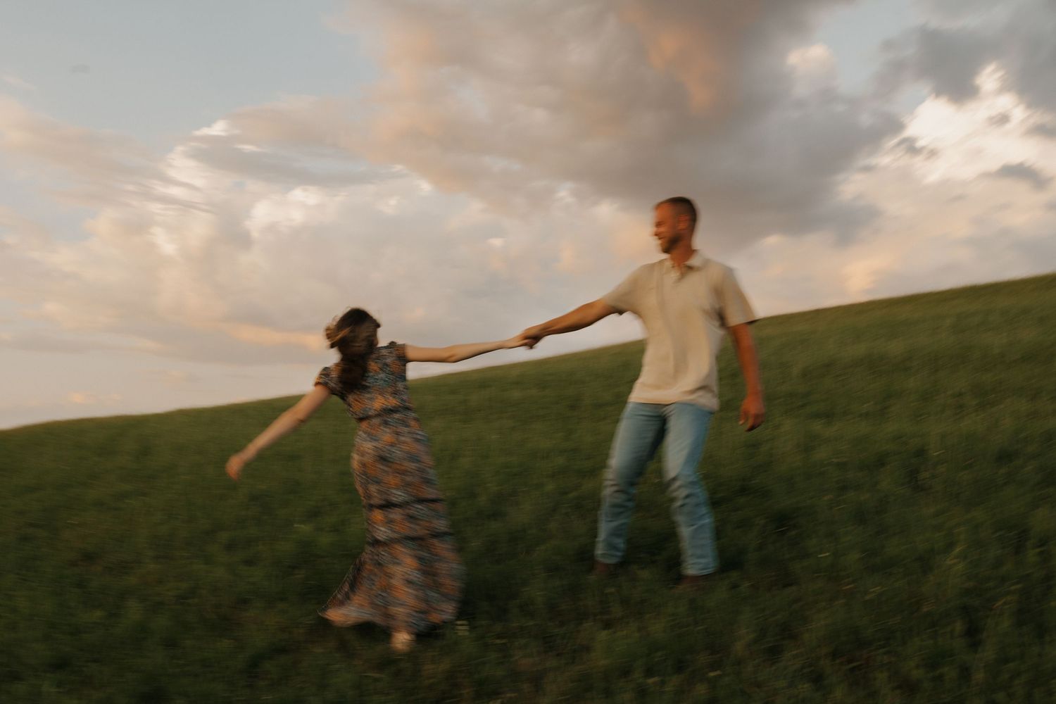 Two people hold hands while standing on a grassy hillside at sunset with warm golden clouds in the sky.