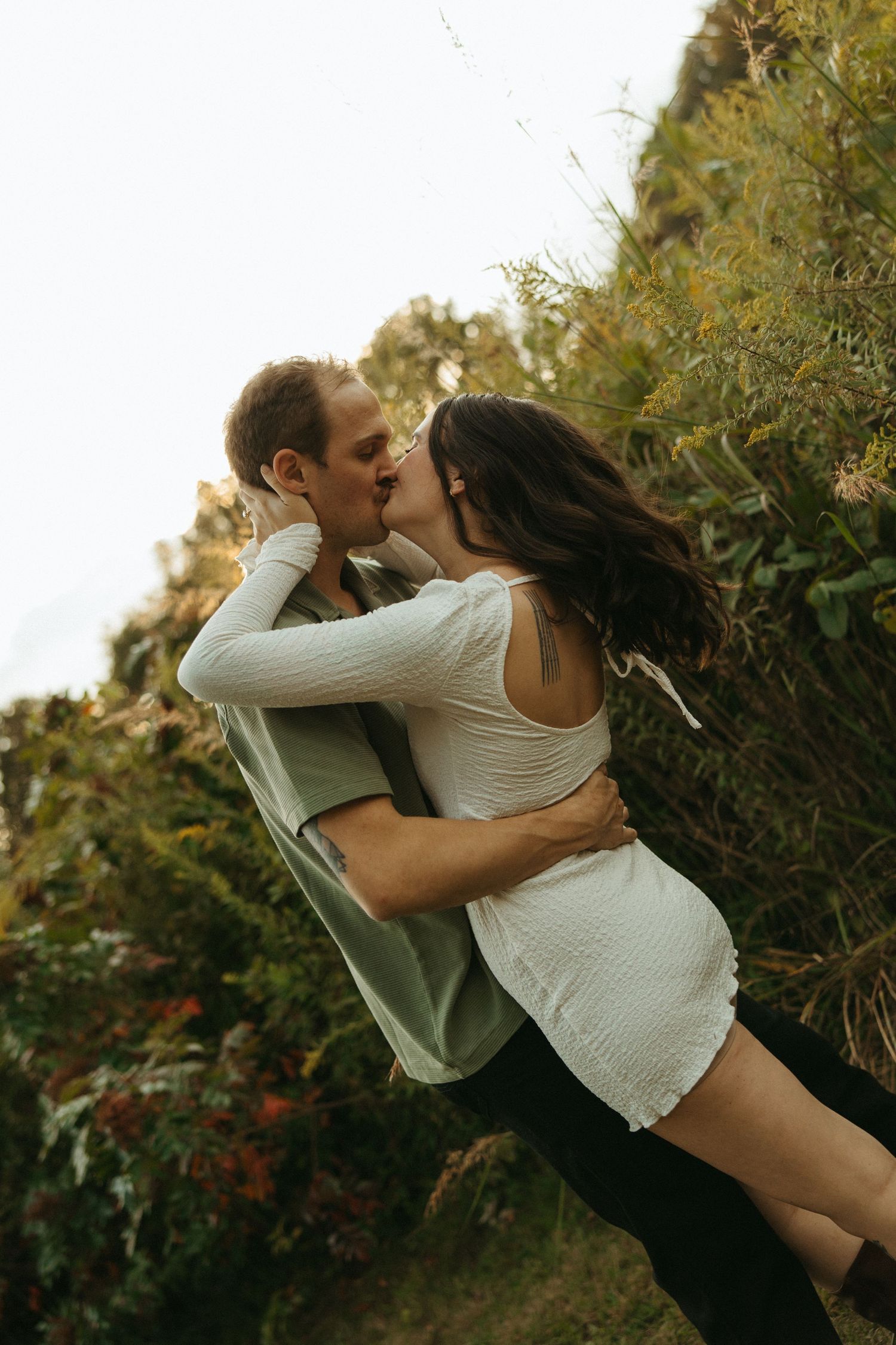A couple sharing a romantic kiss outdoors, embracing among lush green foliage, woman in white dress.