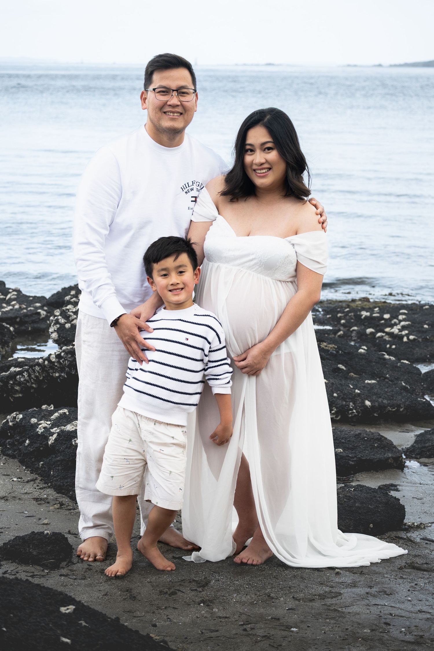 A family wearing coordinated white and striped outfits poses together on a rocky beach at sunset.