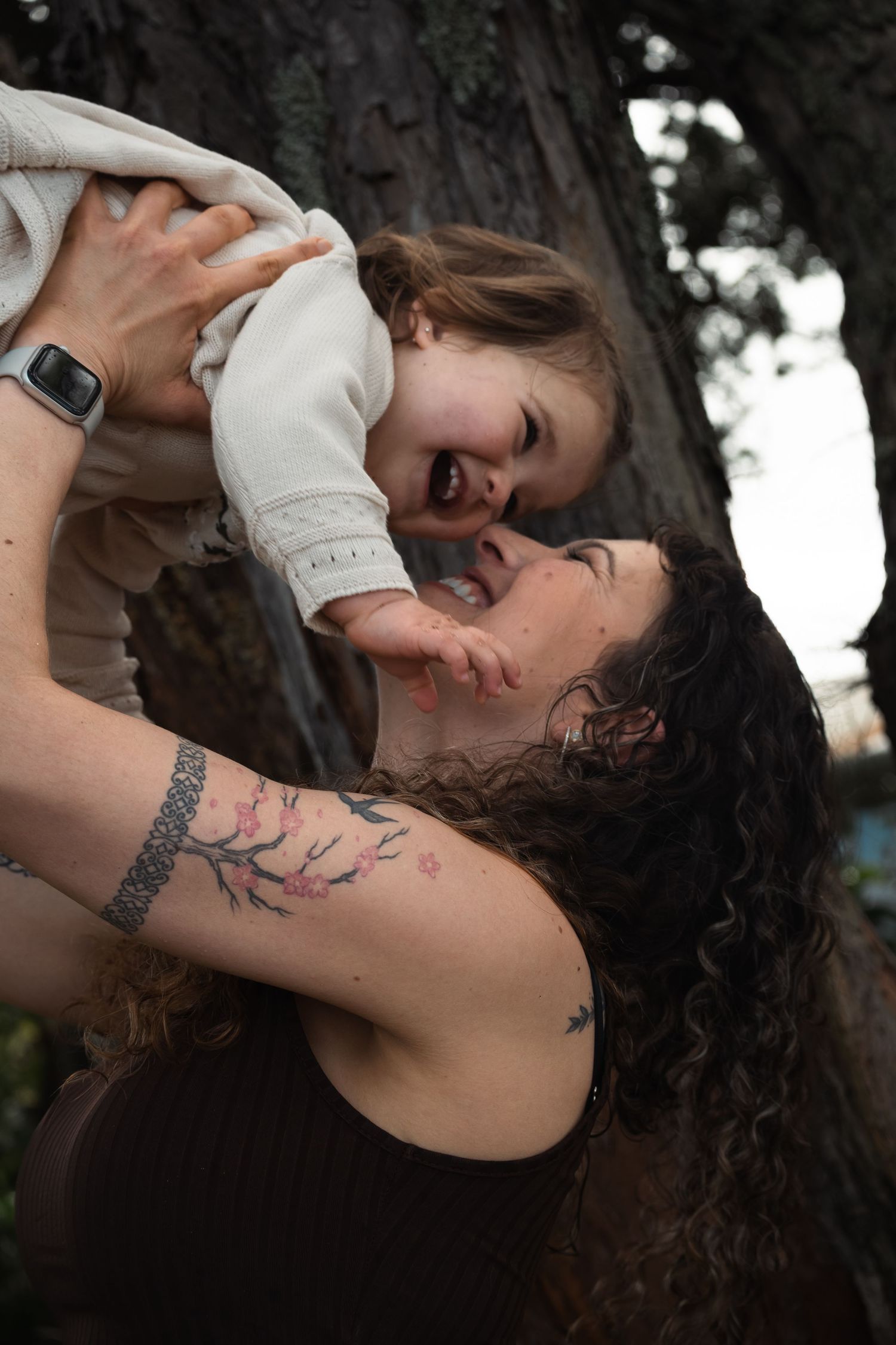 A person with dark curly hair and tattoos leans back against a tree trunk in an artistic outdoor portrait series.