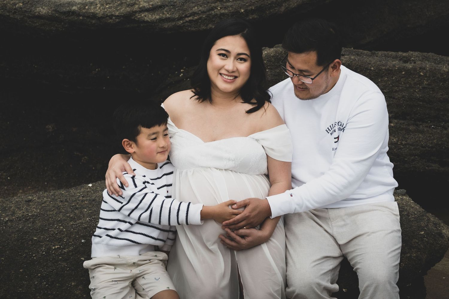 A couple in white outfits shares tender moments during an outdoor photo session against a dark natural backdrop.