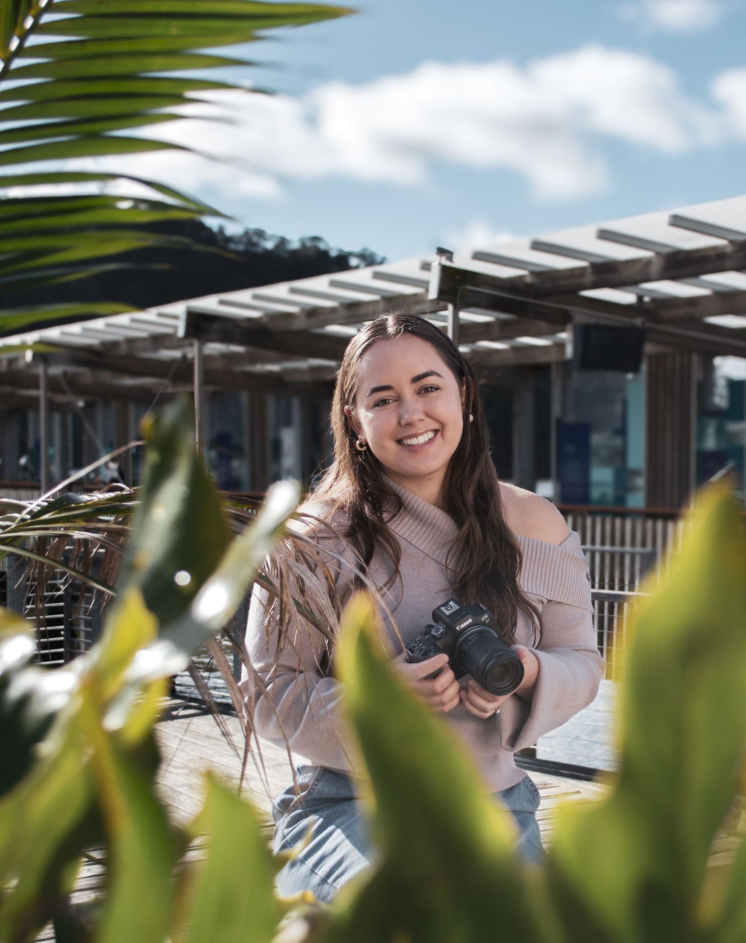 Young woman in casual attire smiles while enjoying outdoor seating area with tropical plants in background.