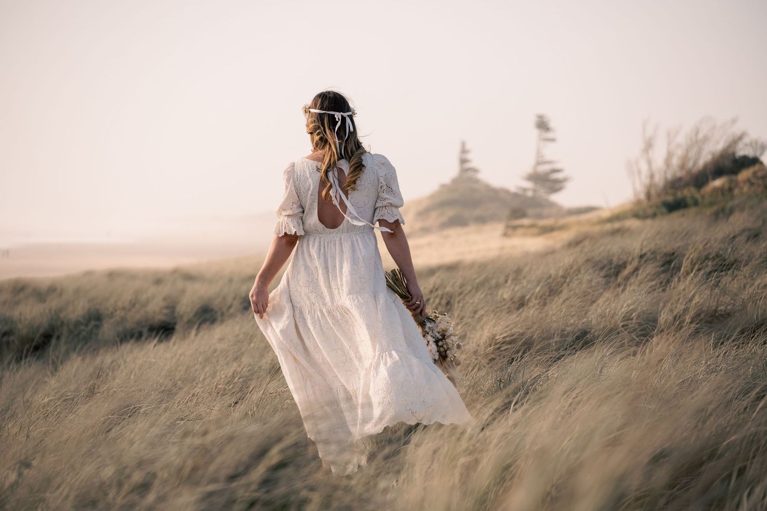 A person in a white dress stands gracefully among tall grass on a windswept coastal hill.