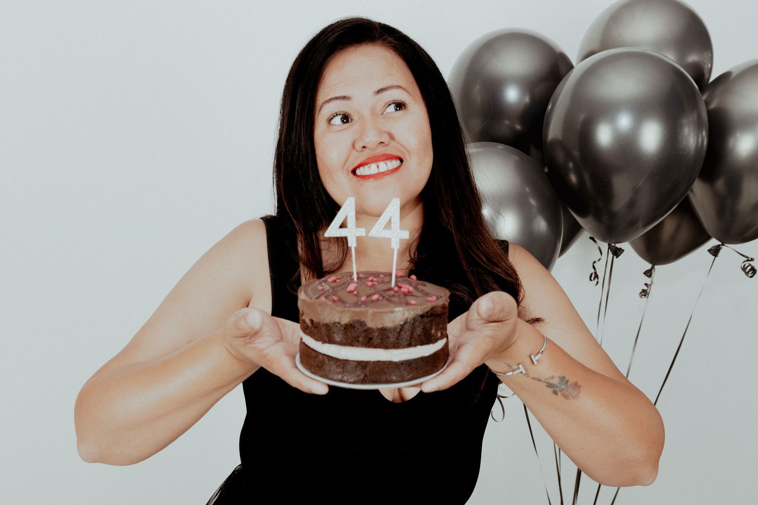 A person celebrates their 44th birthday with black balloons and a chocolate cake with candles.