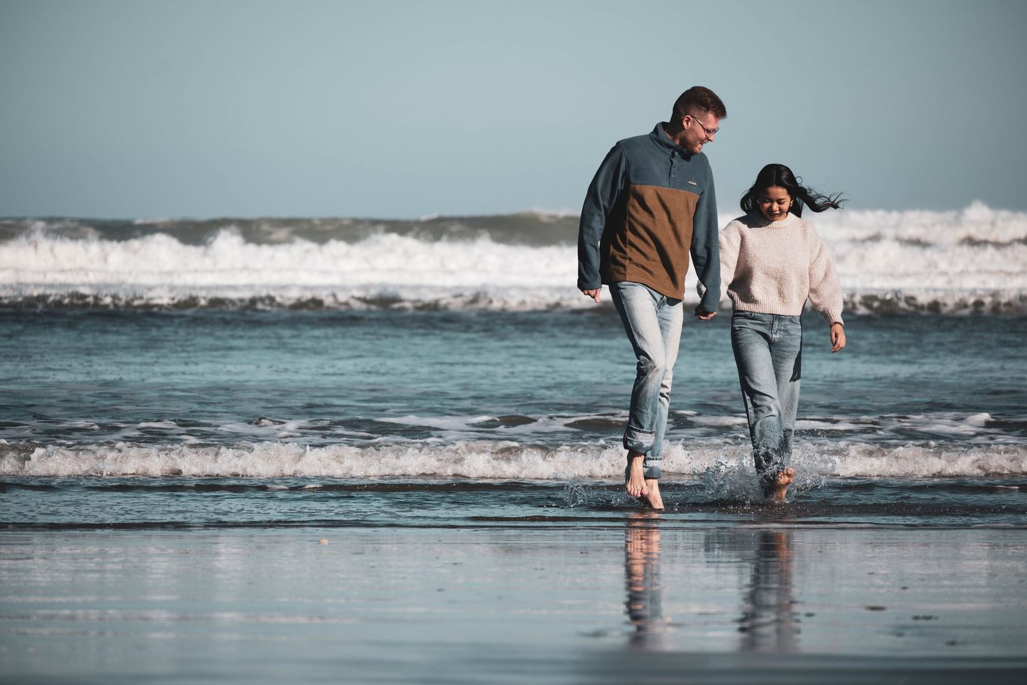 Two people walking along shoreline with waves crashing nearby.