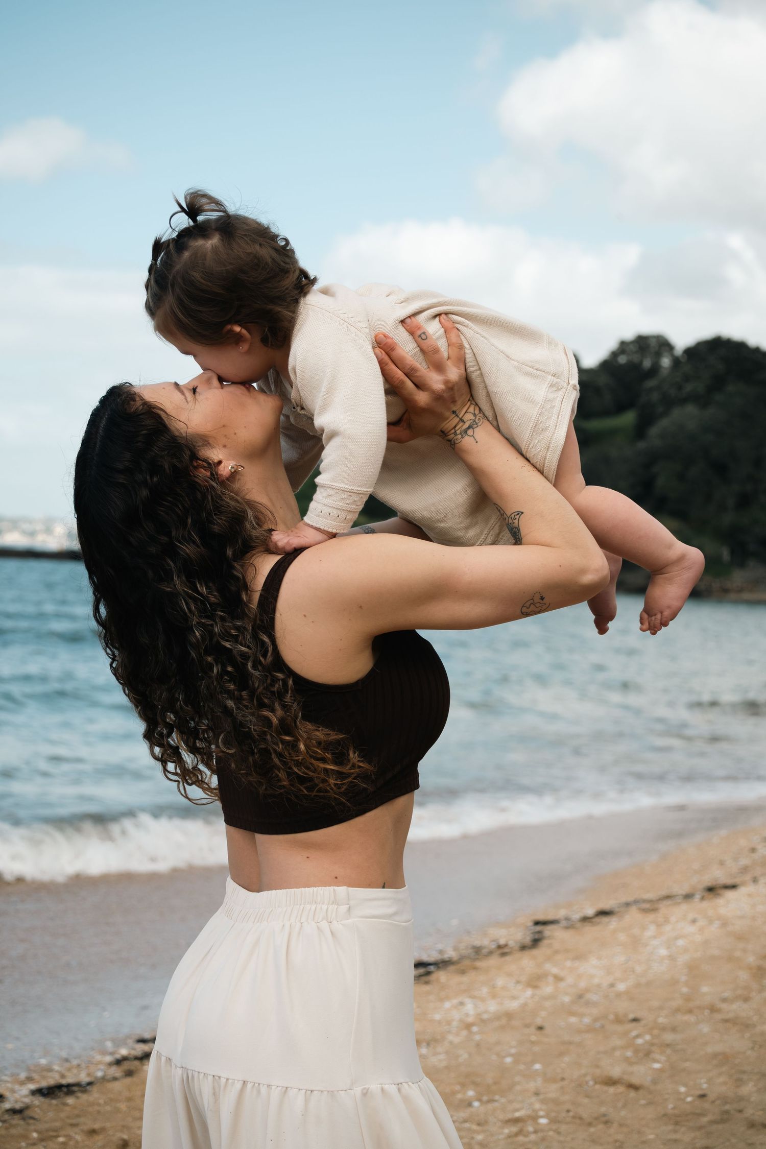 A couple embraces lovingly on a scenic beach with waves and trees in the background.