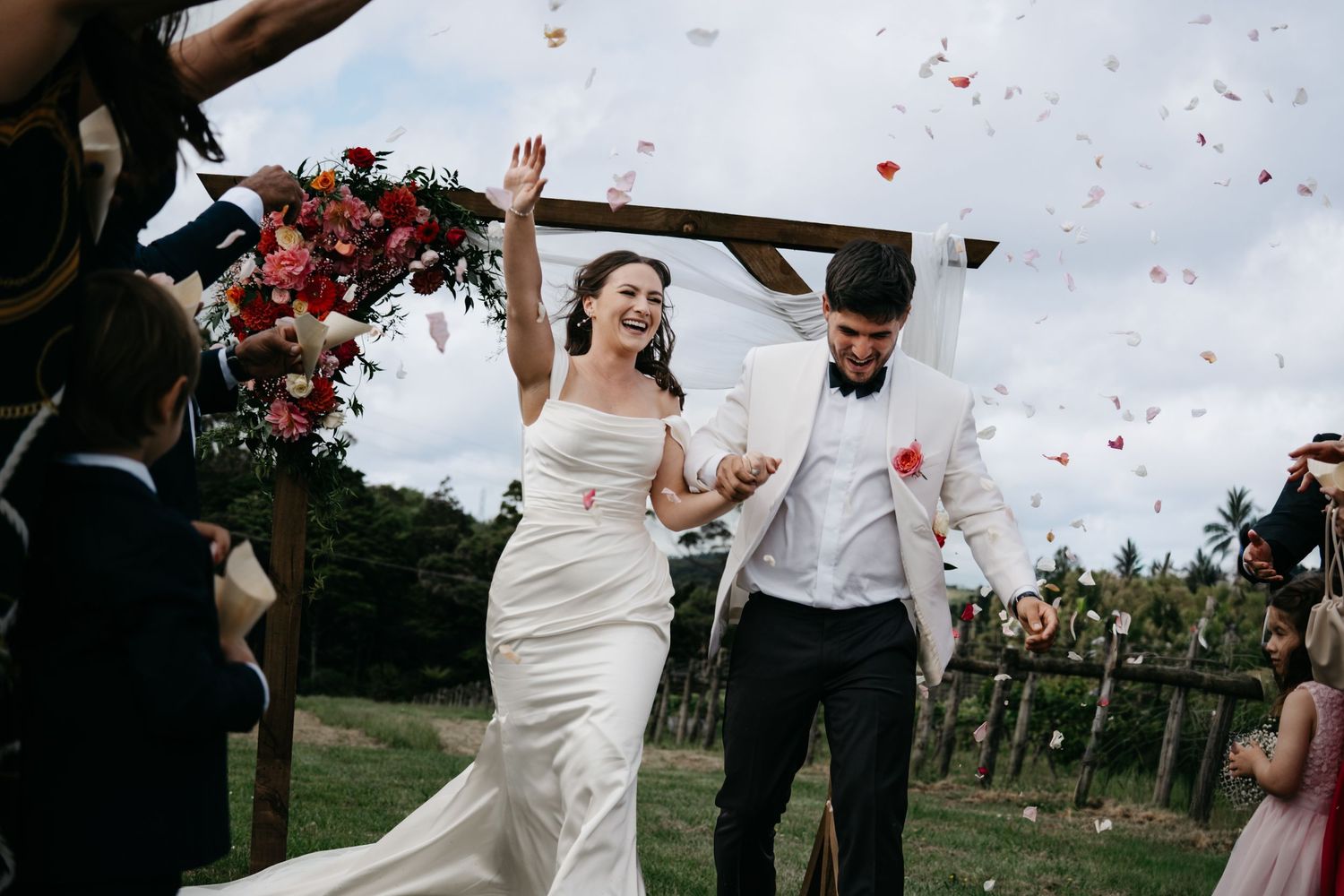 A couple celebrates their wedding day outdoors as red rose petals shower down around them during a joyful moment.