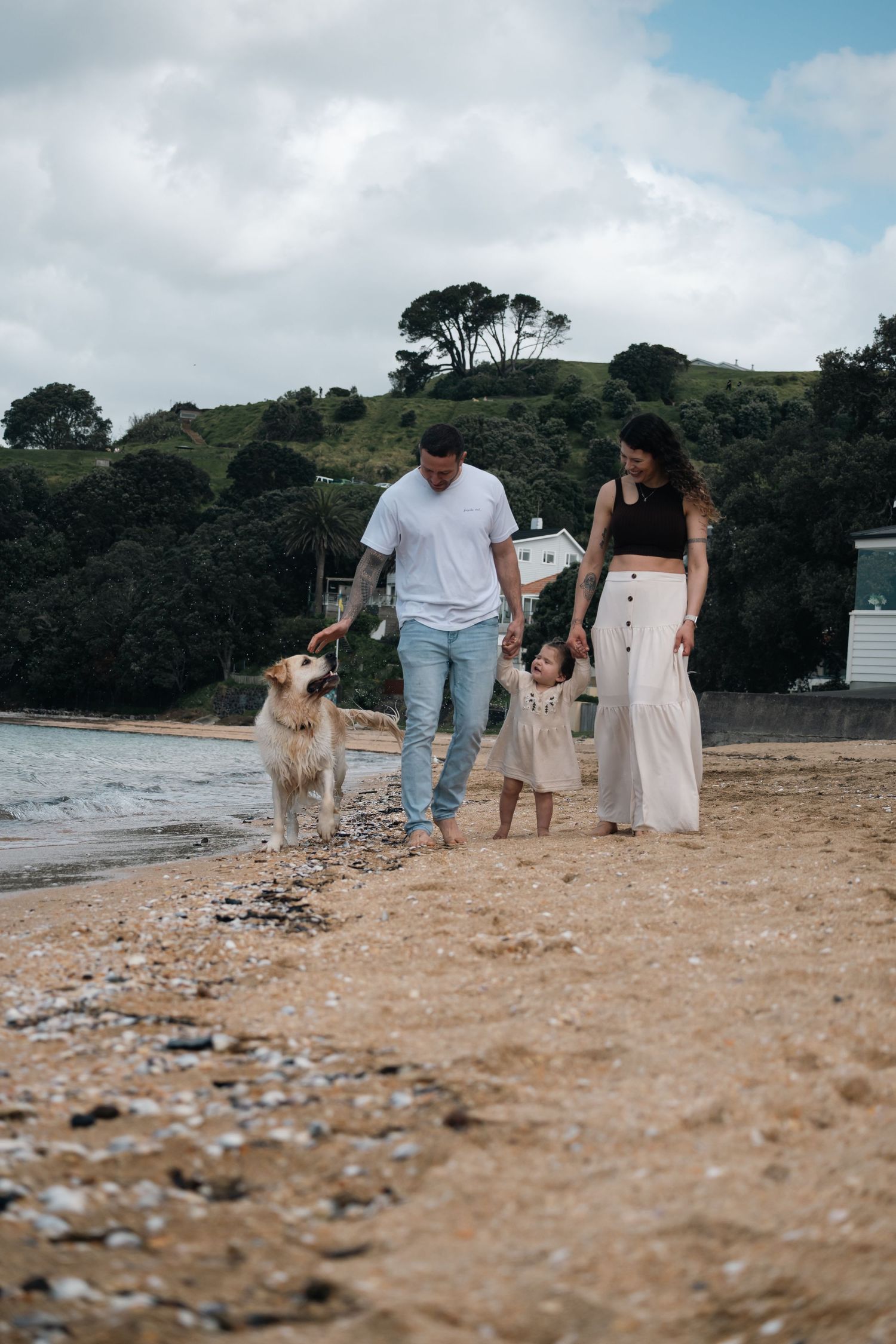 A couple walks with their golden retriever along a rocky beach shoreline on a cloudy day.