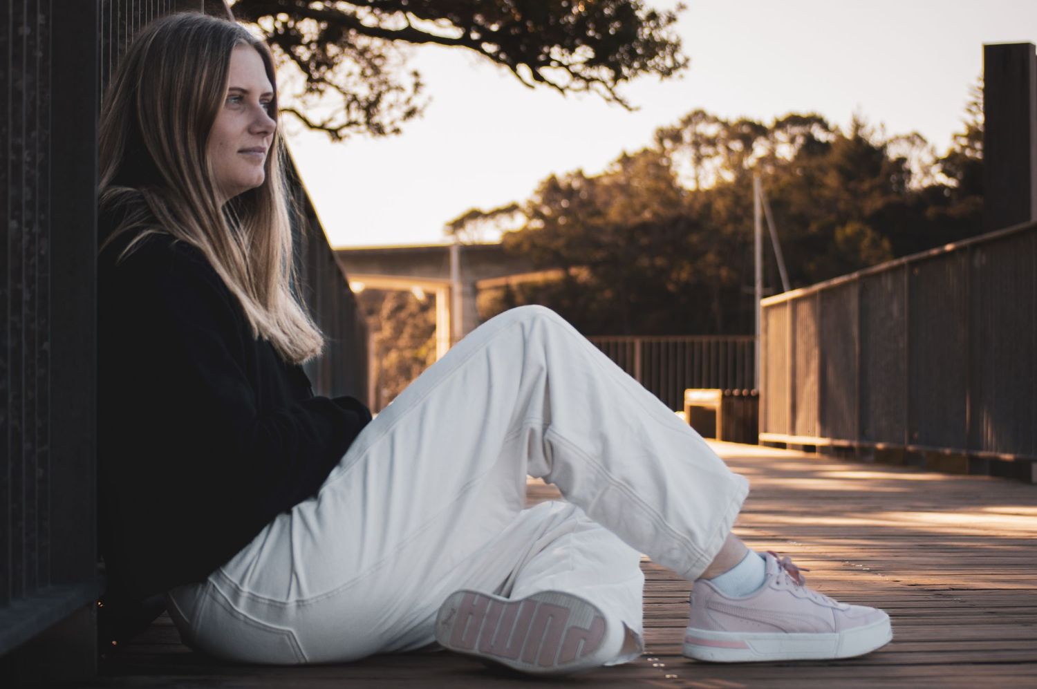 Woman in white sweater and jeans sits on wooden deck during golden hour sunset.