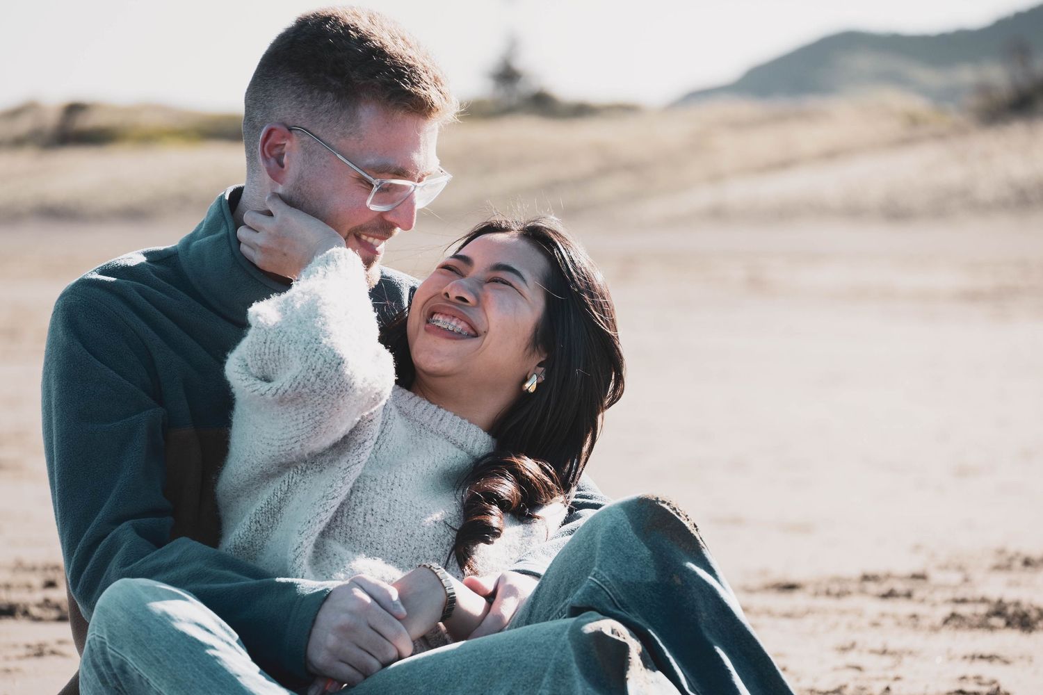 Couple embracing on sandy beach with hills and dunes in background.