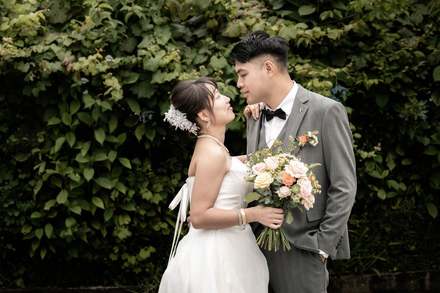 A romantic wedding moment captured outdoors with a beautiful bouquet of peach roses against a lush green hedge backdrop.