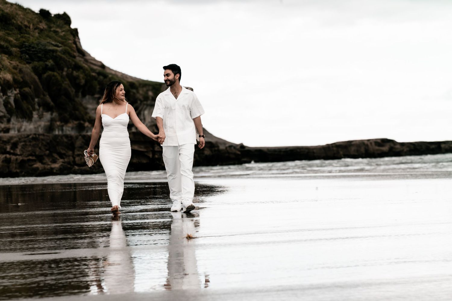 A couple in white attire walking together along a scenic beach shoreline during sunset.
