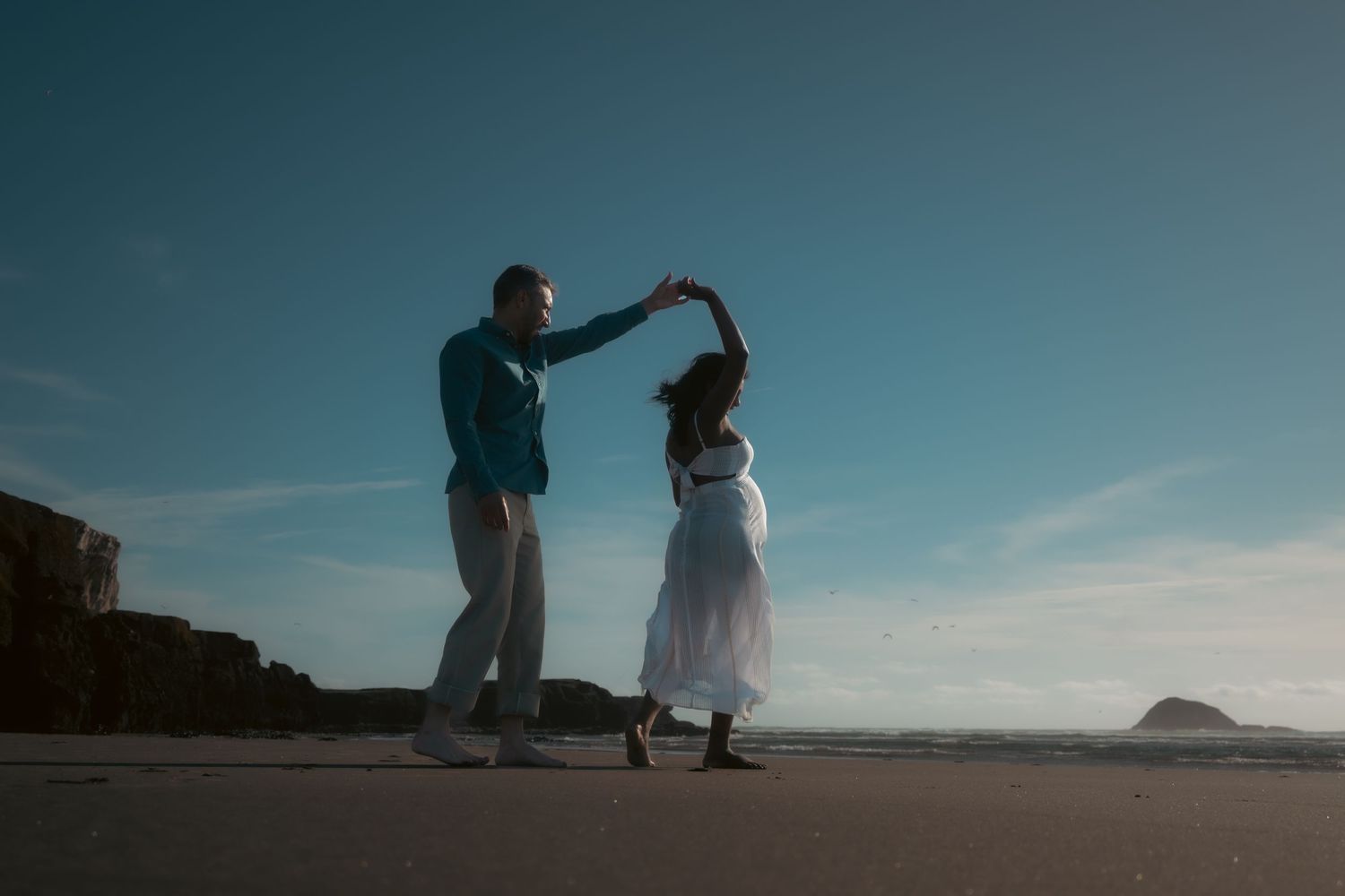 A couple sits on a cliff edge overlooking a scenic coastal landscape at sunset.
