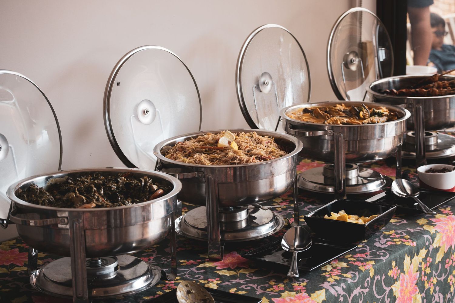 A row of stainless steel chafing dishes with glass lids on a floral tablecloth at a buffet setup.