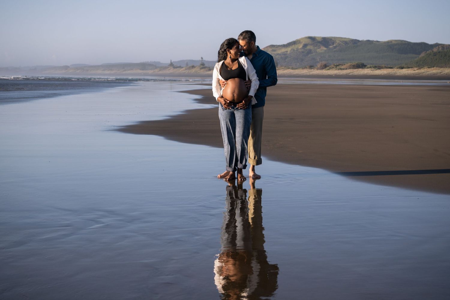 A couple embraces on a reflective beach at sunset, their silhouette mirrored in the wet sand along the shoreline.