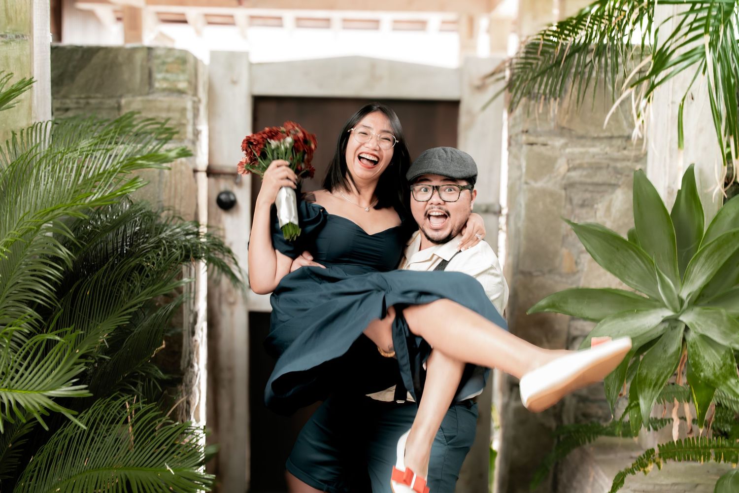 A couple shares joyful moments together in a garden setting with stone walls and tropical plants in the background.