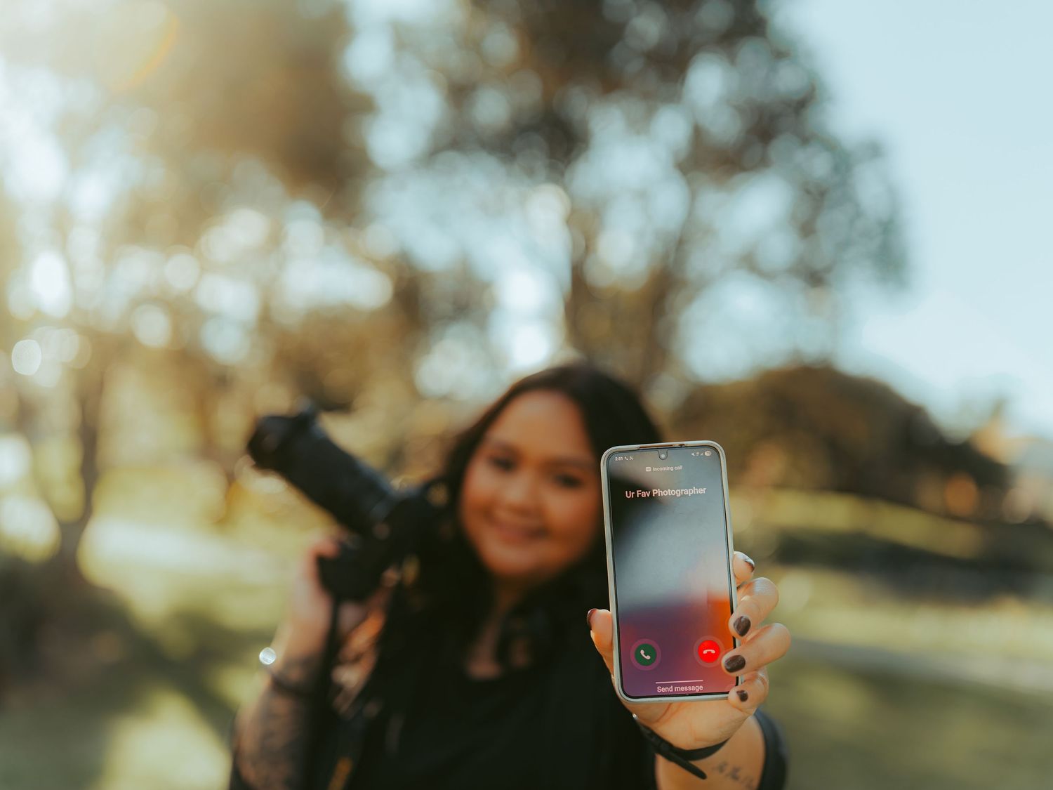 Woman holding smartphone toward camera with incoming call, carrying DSLR camera outdoors in golden sunlight.