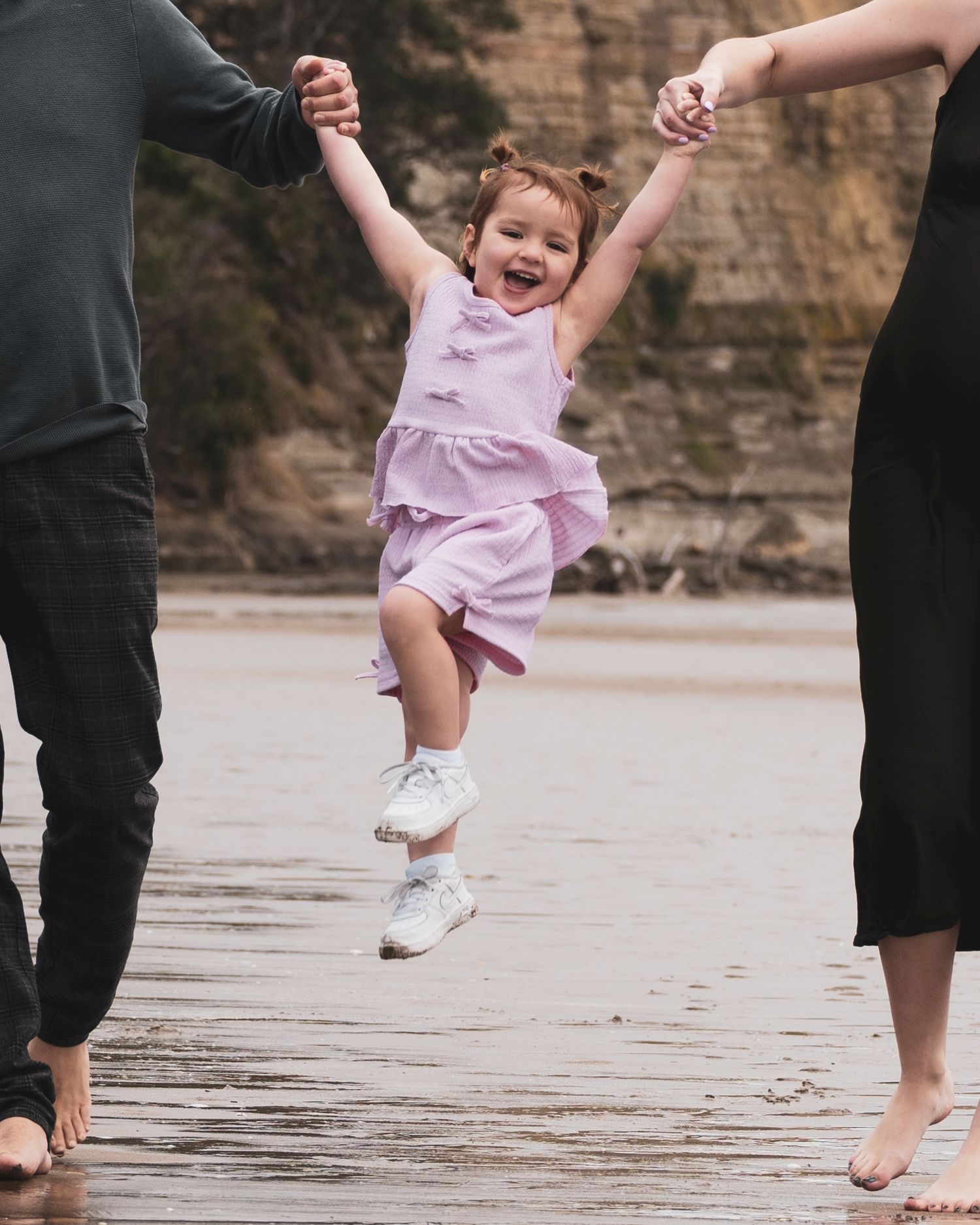 A joyful child in pink clothing jumps while being held by adults' hands on a beach.