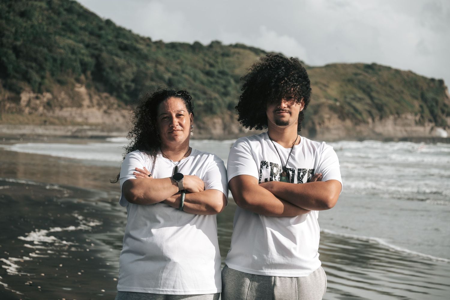 Two people in white t-shirts stand with crossed arms on a scenic beach with cliffs and waves in the background.