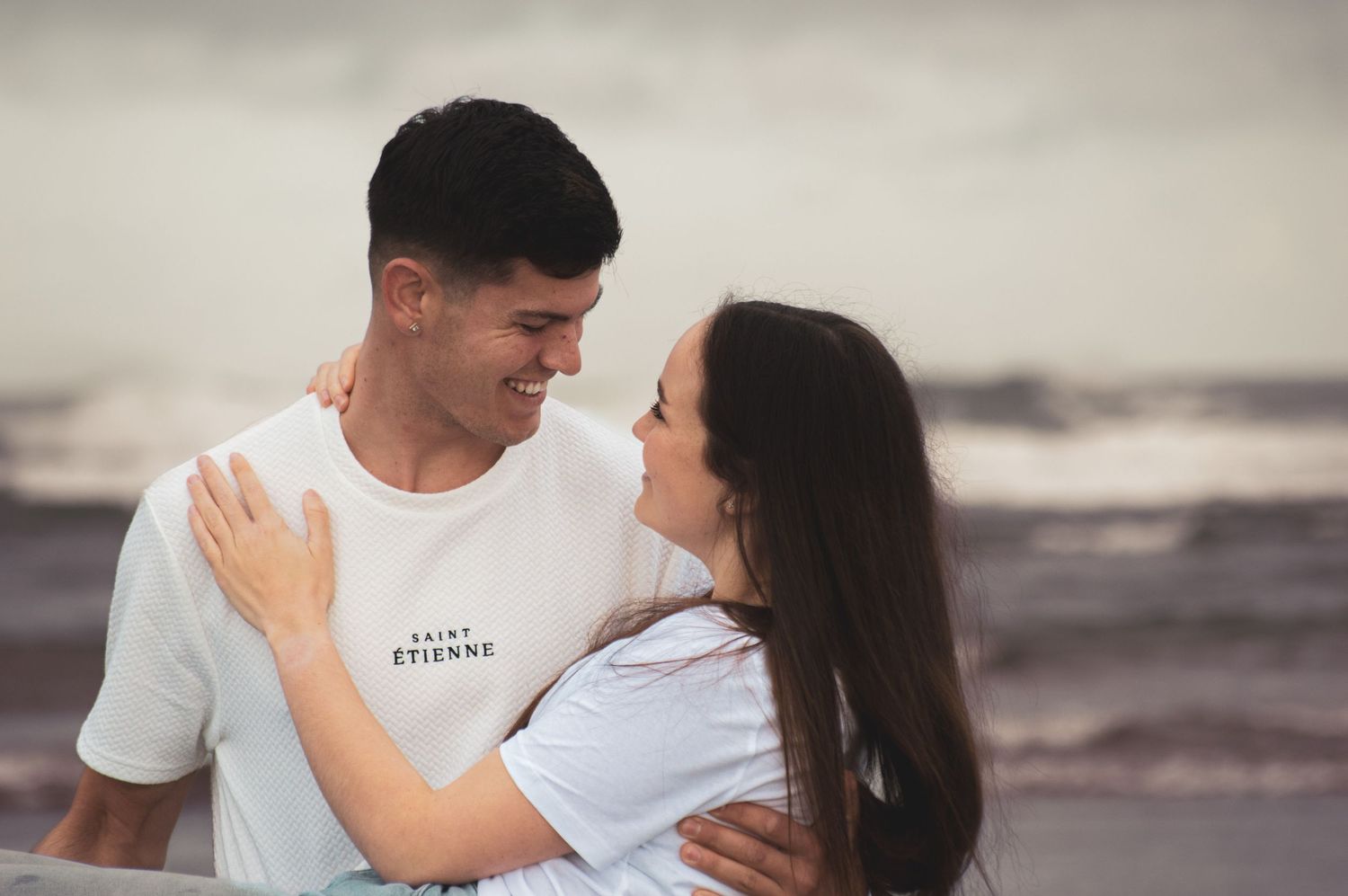 Couple embracing on a cloudy beach wearing white and grey casual clothing.