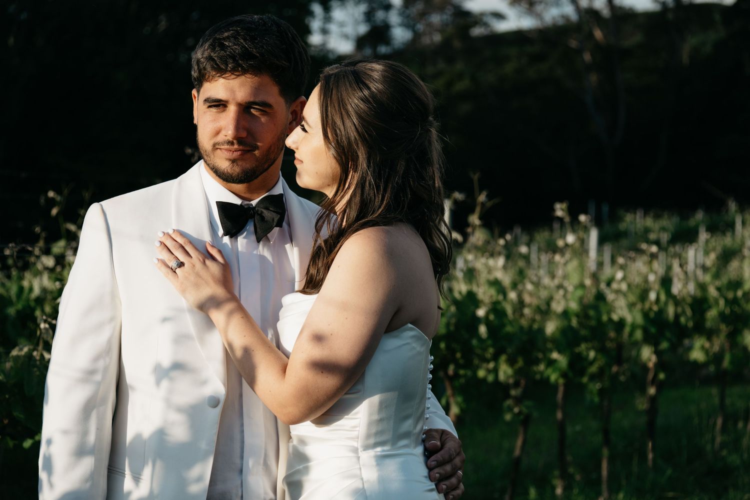 A couple in wedding attire embraces in a sunlit garden with wildflowers in the background.