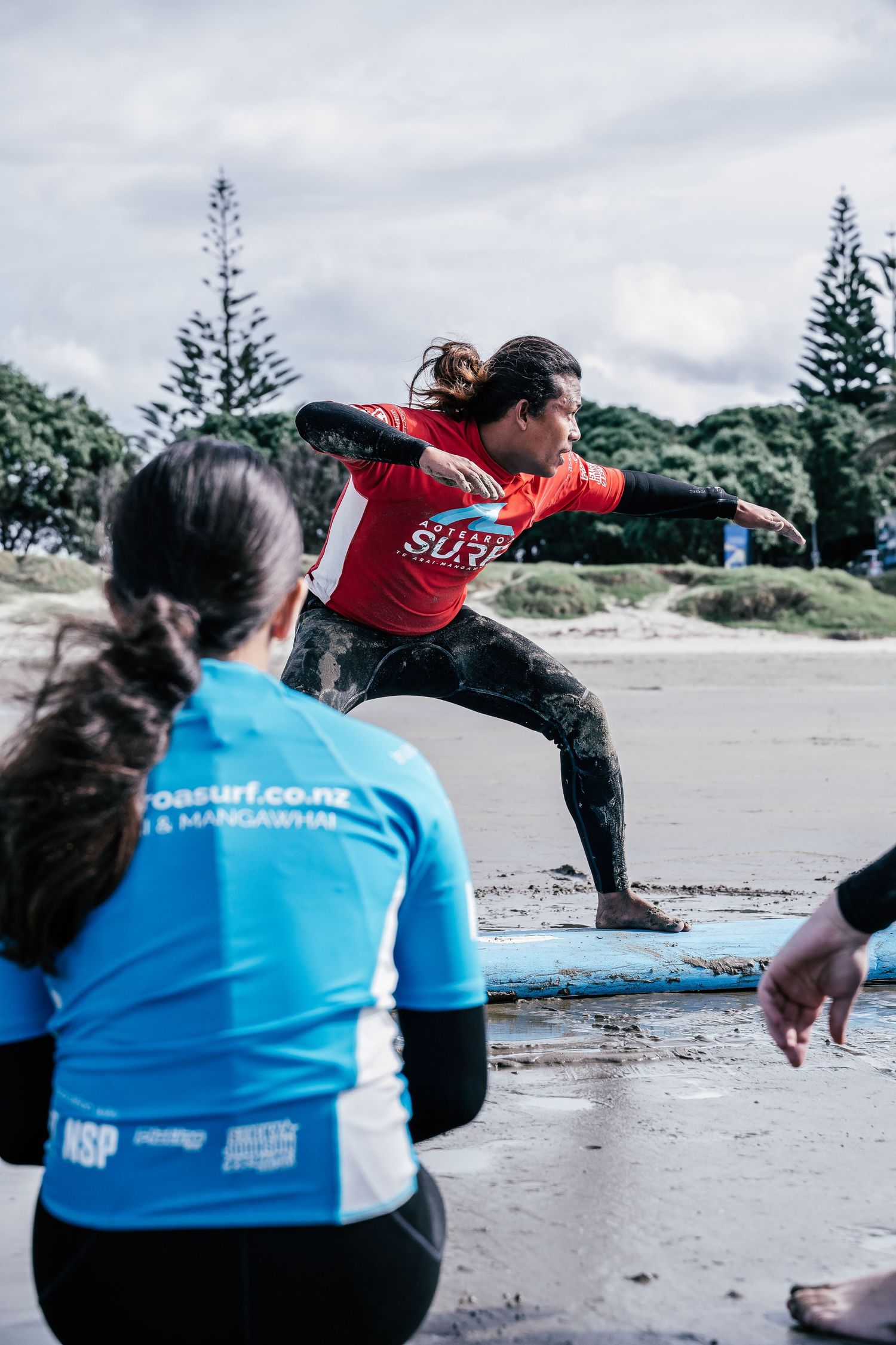 Surf instructor in red shirt demonstrates surfing stance on beach while student in blue rash guard watches during lesson in New Zealand.