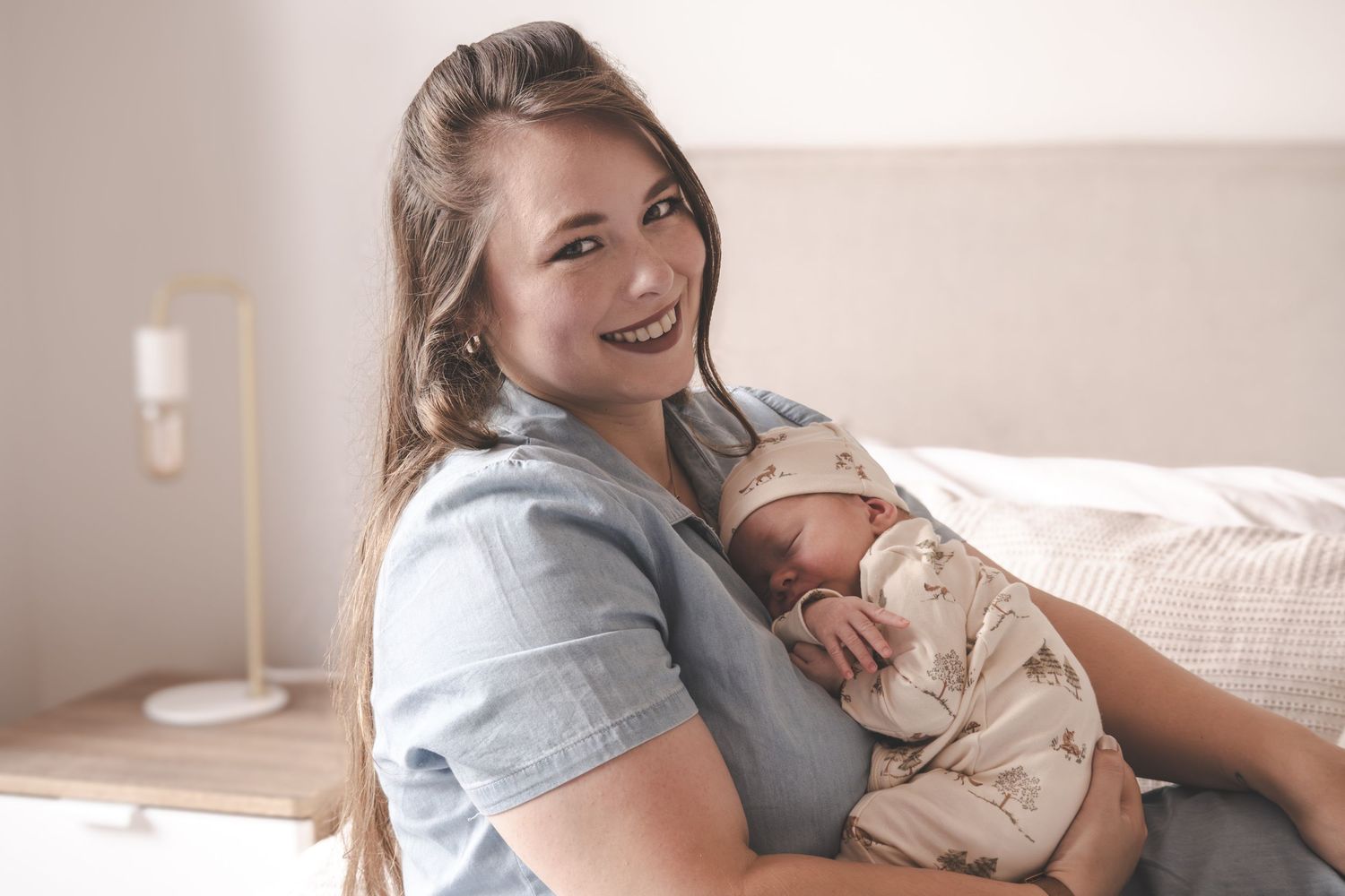A person in a gray shirt holds and smiles at a sleeping newborn baby in a cozy bedroom setting.
